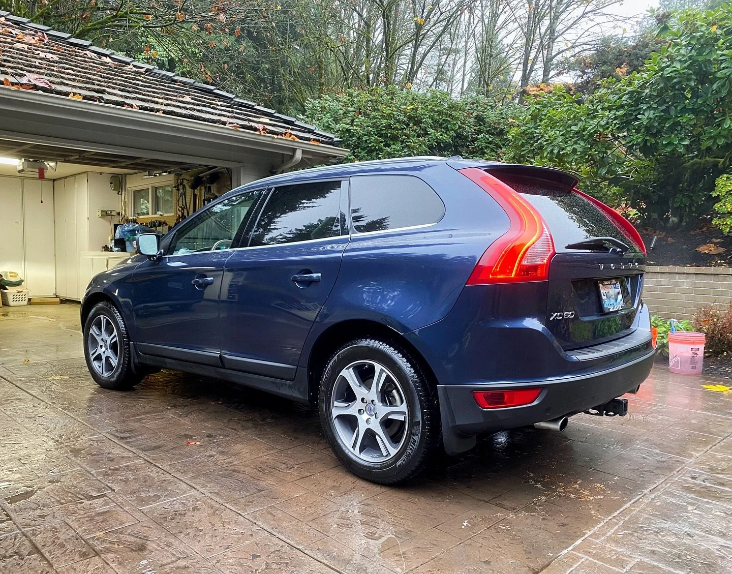 Blue Volvo XC60 parked on a wet driveway in a residential area with green trees and bushes in the background, and a garage visible on the left side.