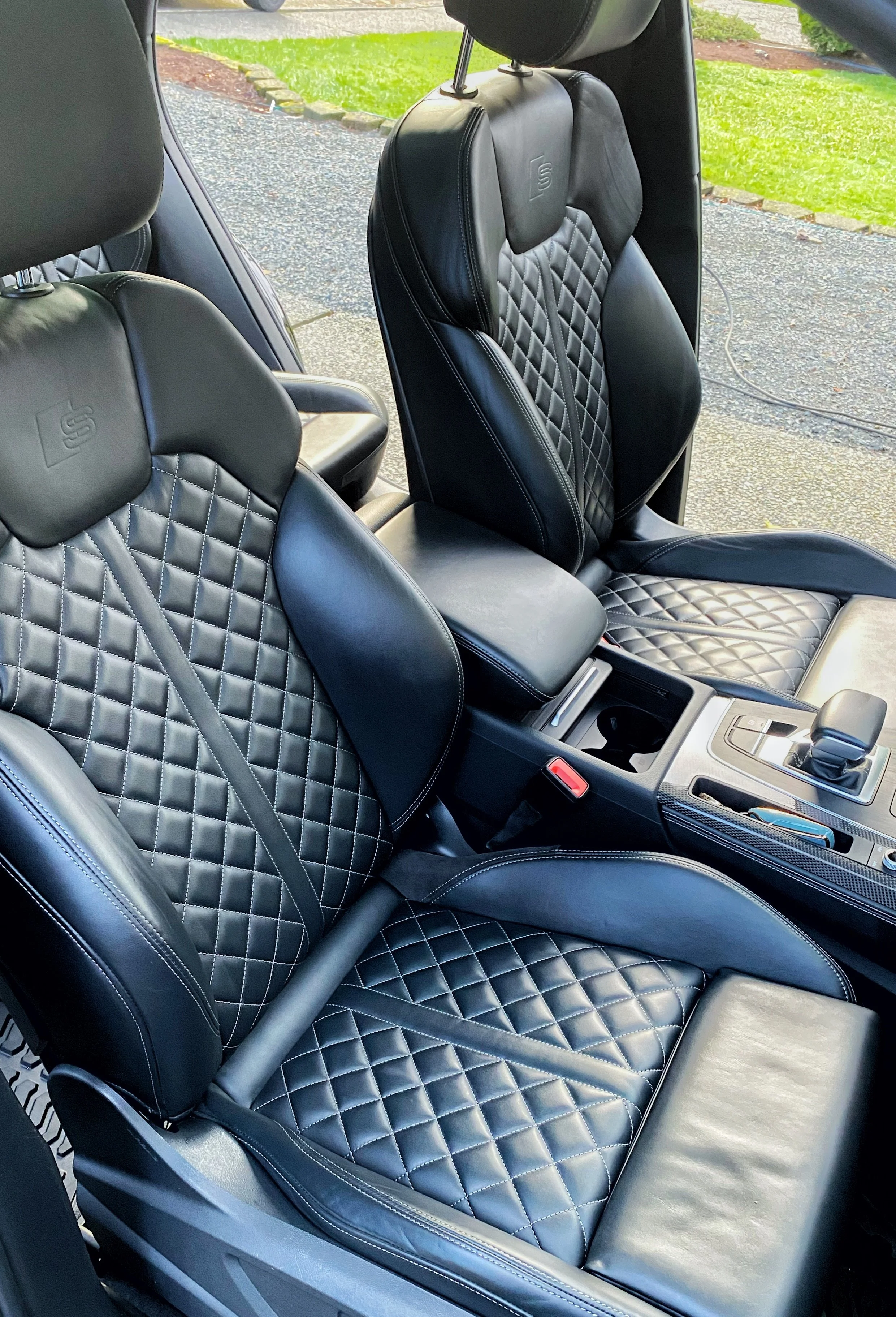 The interior of a car showing two black leather seats with diamond-stitched pattern and headrests, part of the center console with gear shift, and a view of a driveway and grass outside the window. 