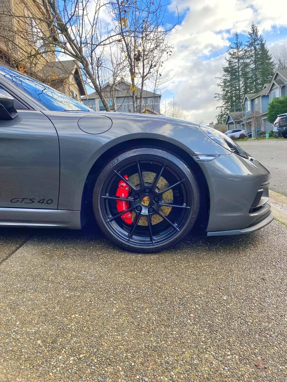 Close-up of a gray Porsche GTS 4.0 sports car parked on a street, showing the front wheel with a red brake caliper and a yellow brake disc, along with the house and trees in the background.