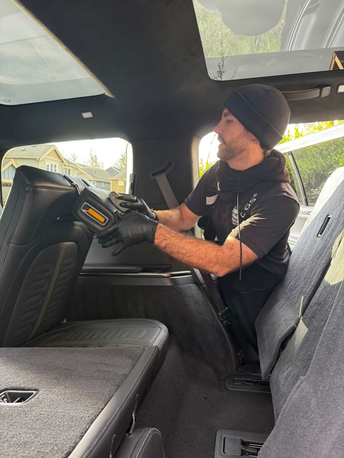 A man wearing a black knit cap, black gloves, and a black jacket with a patch, is working inside the back of a vehicle, likely a police or emergency vehicle, while standing in the rear area.