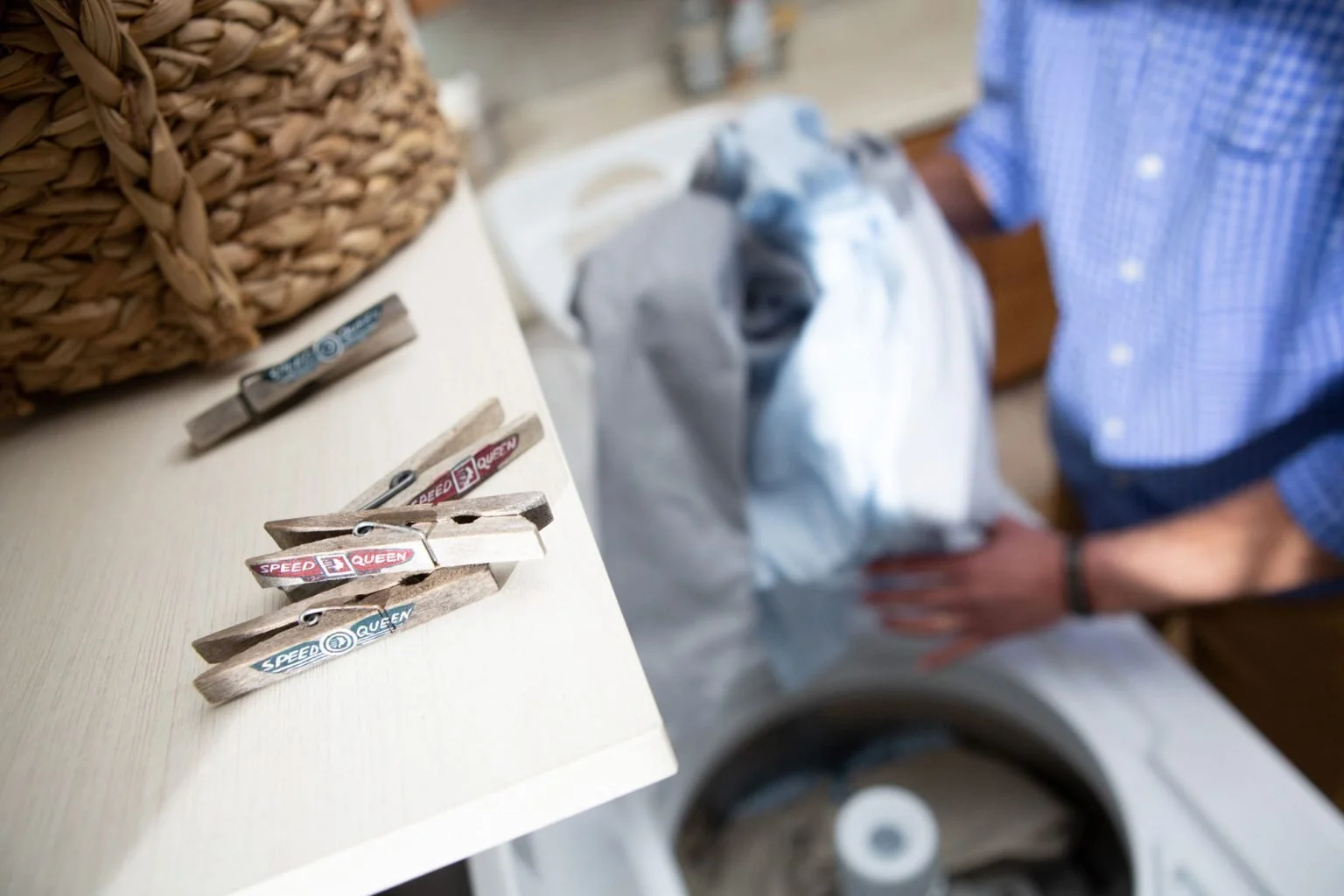 Close-up of small wooden clothespins on a white surface, with a person in a blue checked shirt in the background near a laundry basket and a washing machine.