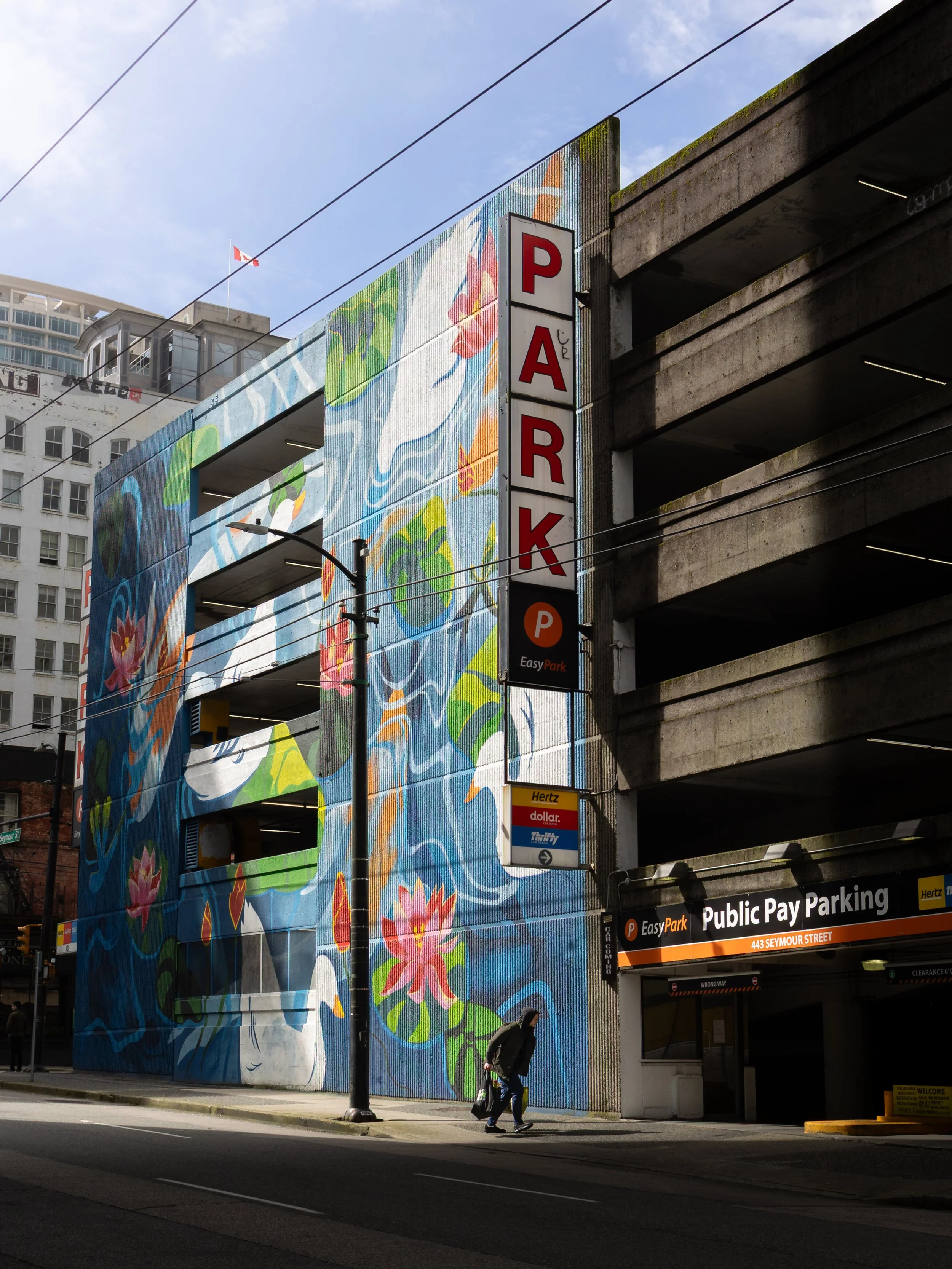 Multilevel parking garage with colorful murals on the exterior wall, a vertical parking sign, and a homeless person walking on the sidewalk carrying a bag.
