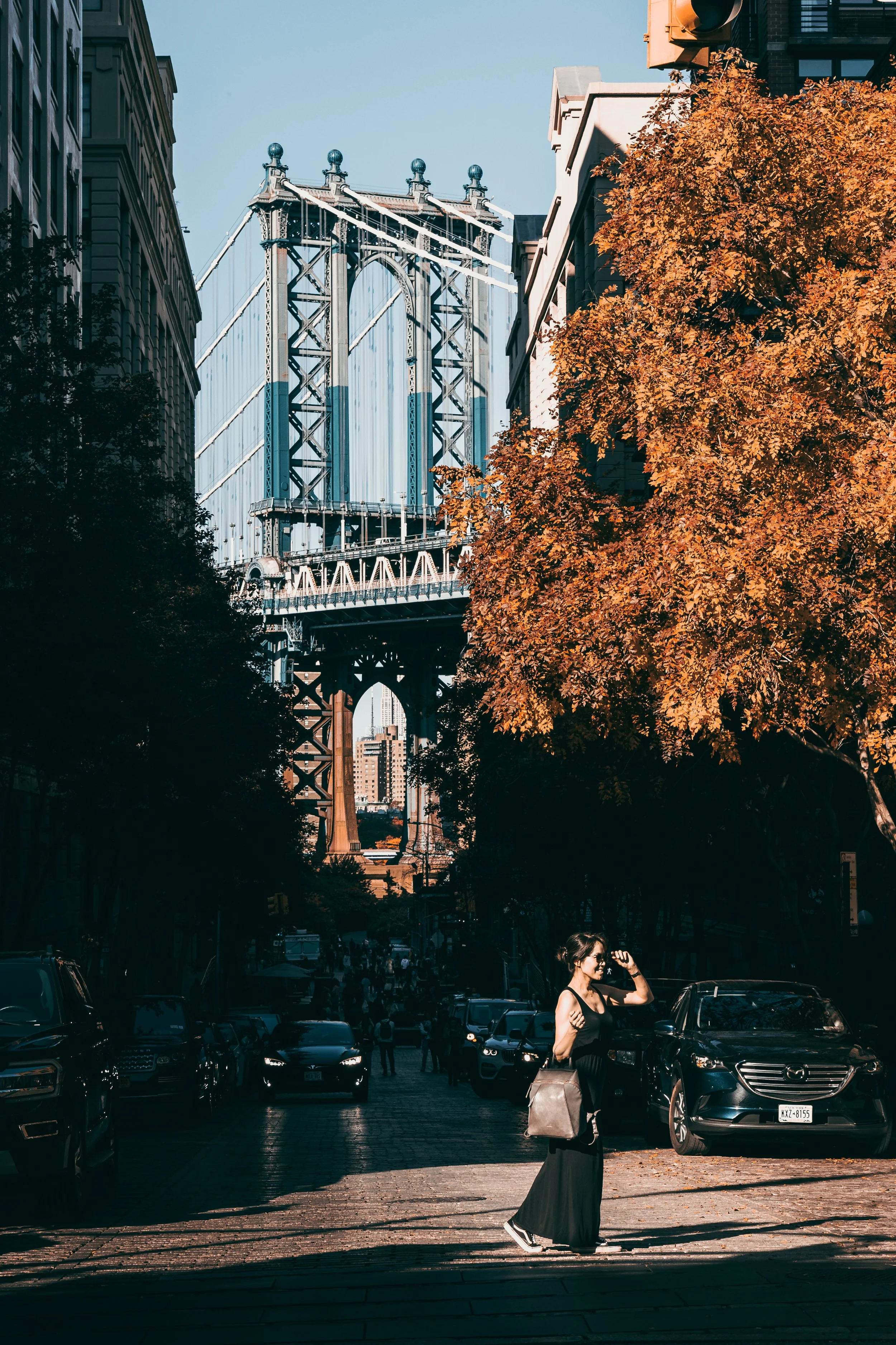 A woman walking on a city street with cars parked on the side and a large orange tree, with the Manhattan Bridge in the background in New York City.