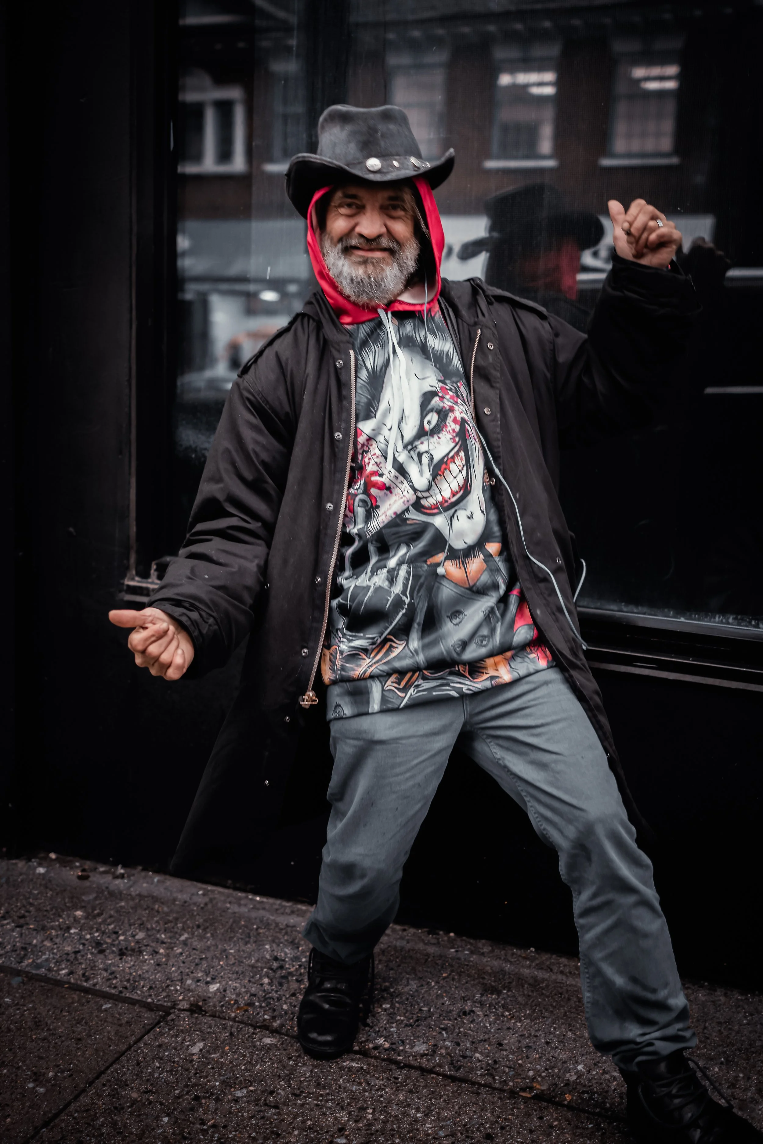 A man with a gray beard wearing a black cowboy hat, black jacket, graphic T-shirt, gray pants, and black shoes, standing outdoors on a wet sidewalk and posing with an enthusiastic stance in front of a black window.