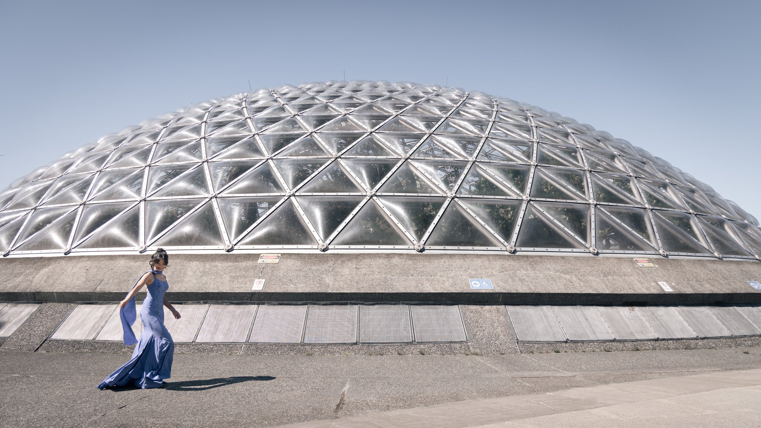 A young woman in a long flowy blue dress walking in front of a large glass geodesic dome structure known as the Bloedel Observatory in Vancouver, BC, Canada.