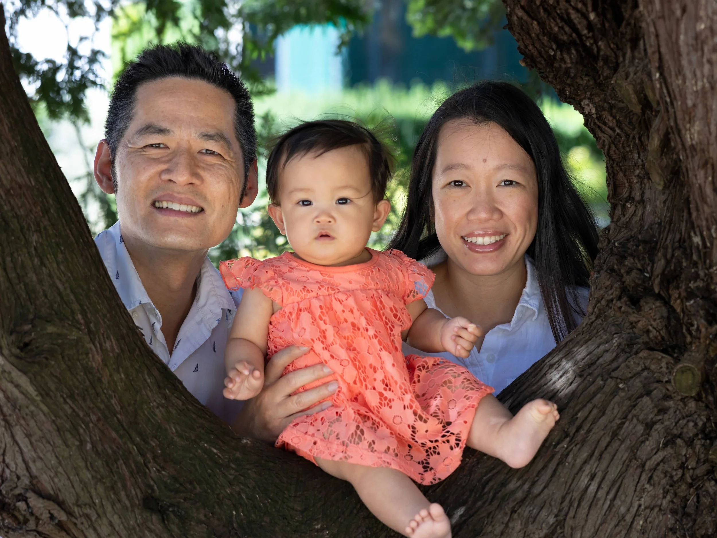 A family of three, a man, a woman, and a baby girl, sitting inside a tree hollow outdoors with green foliage in the background, smiling at the camera.