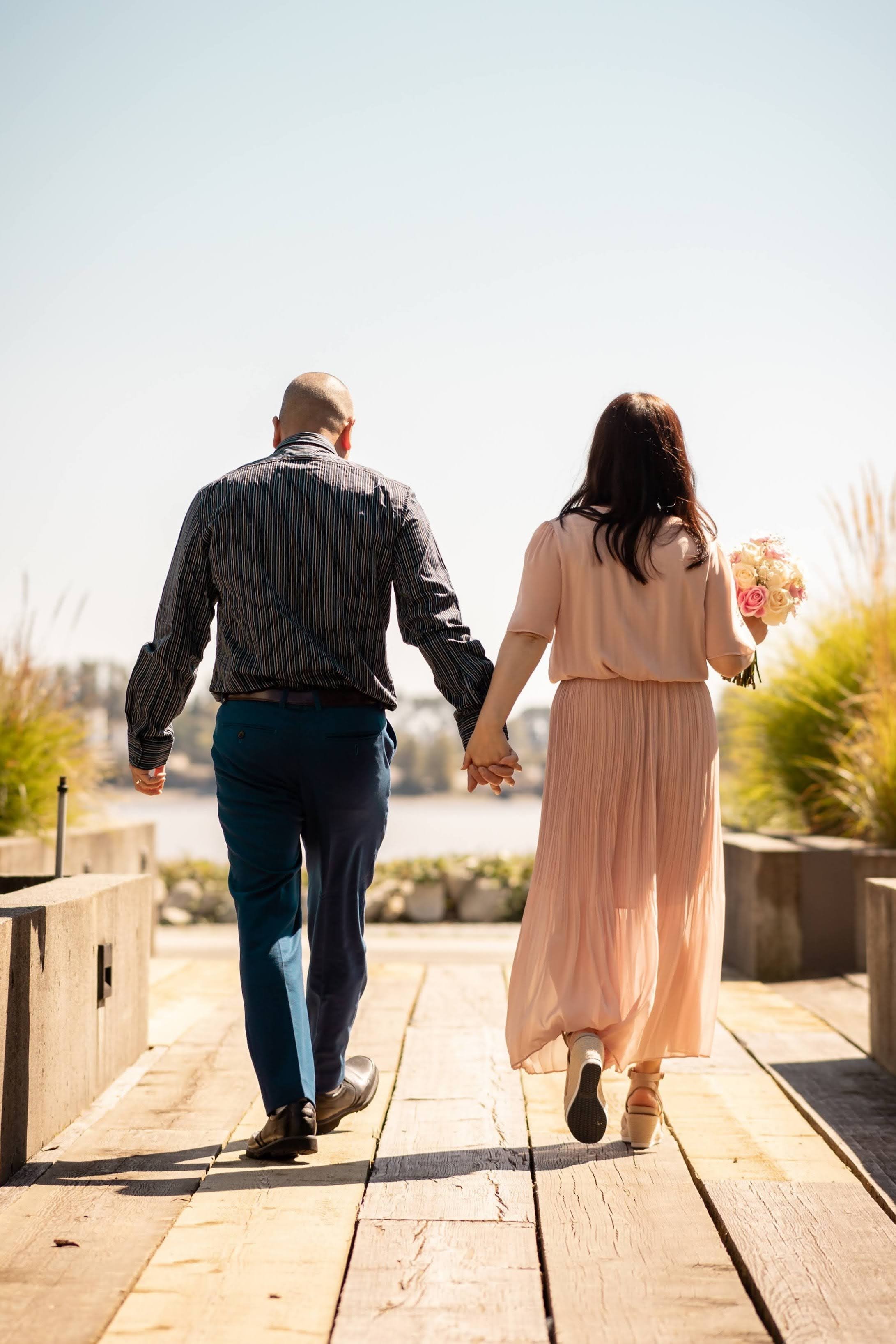 A couple holding hands while walking on a wooden path outdoors, with water and greenery in the background. The woman is holding a bouquet of flowers.