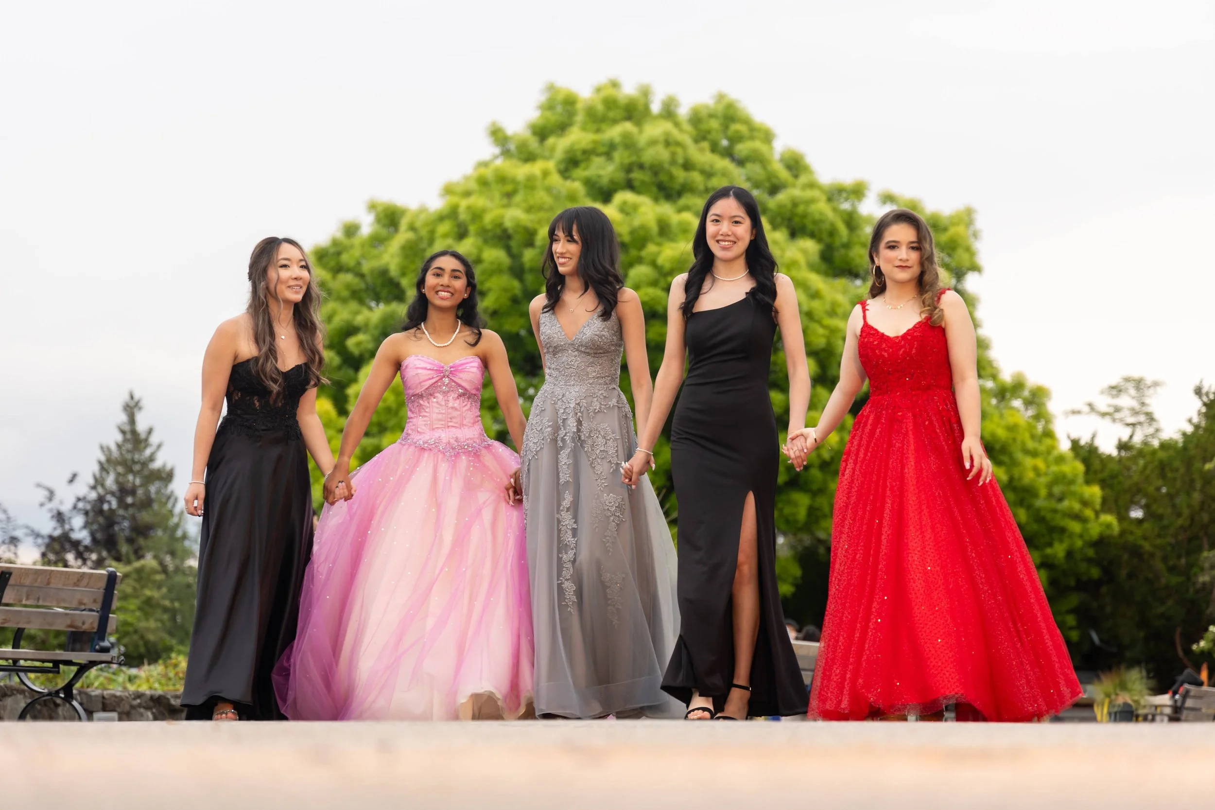 Group of five young women in formal prom dresses holding hands and walking outdoors in front of a large green tree.