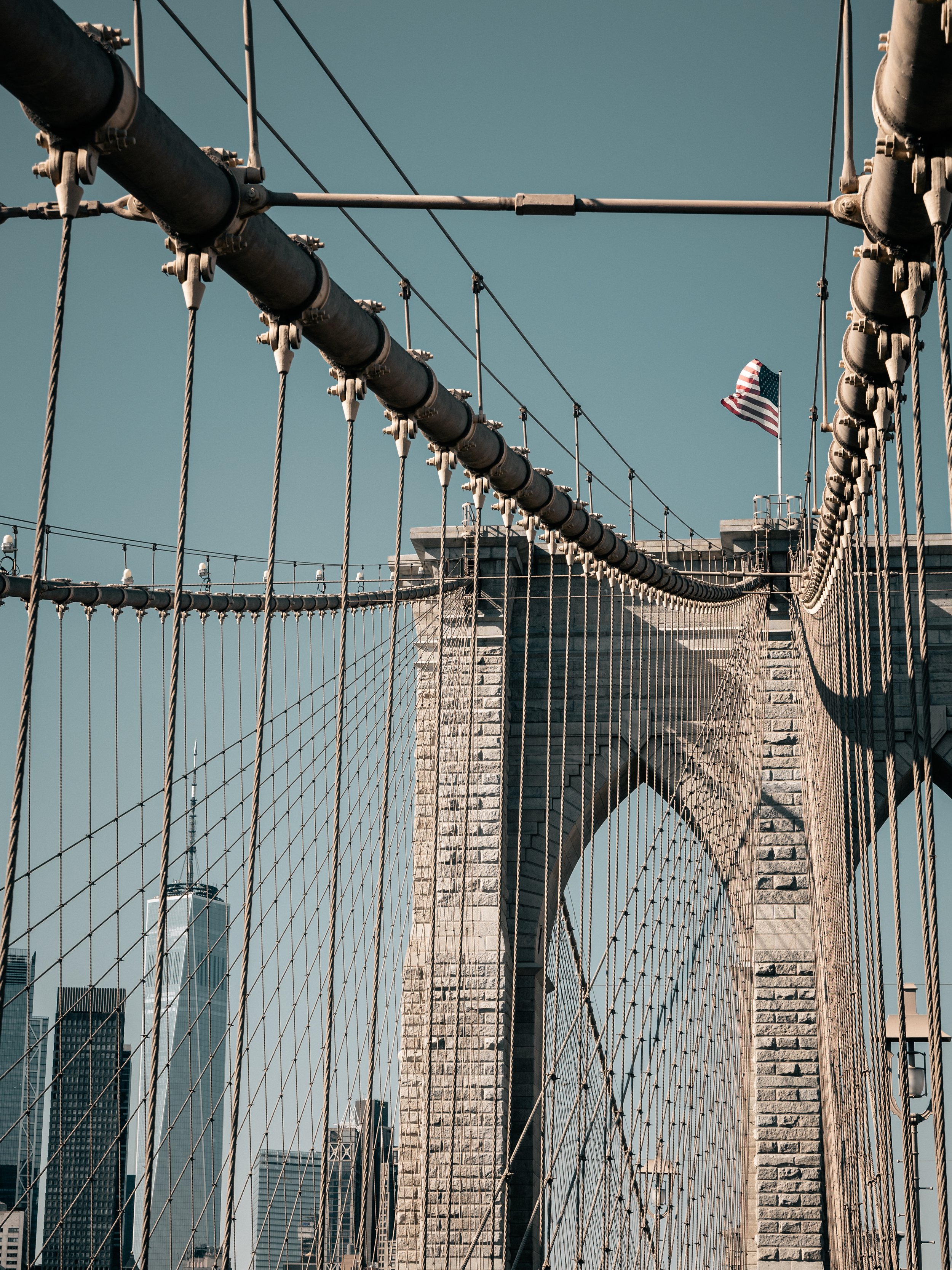 Looking up at the Brooklyn Bridge in New York City, with the American flag flying on top of the bridge and city skyscrapers in the background.