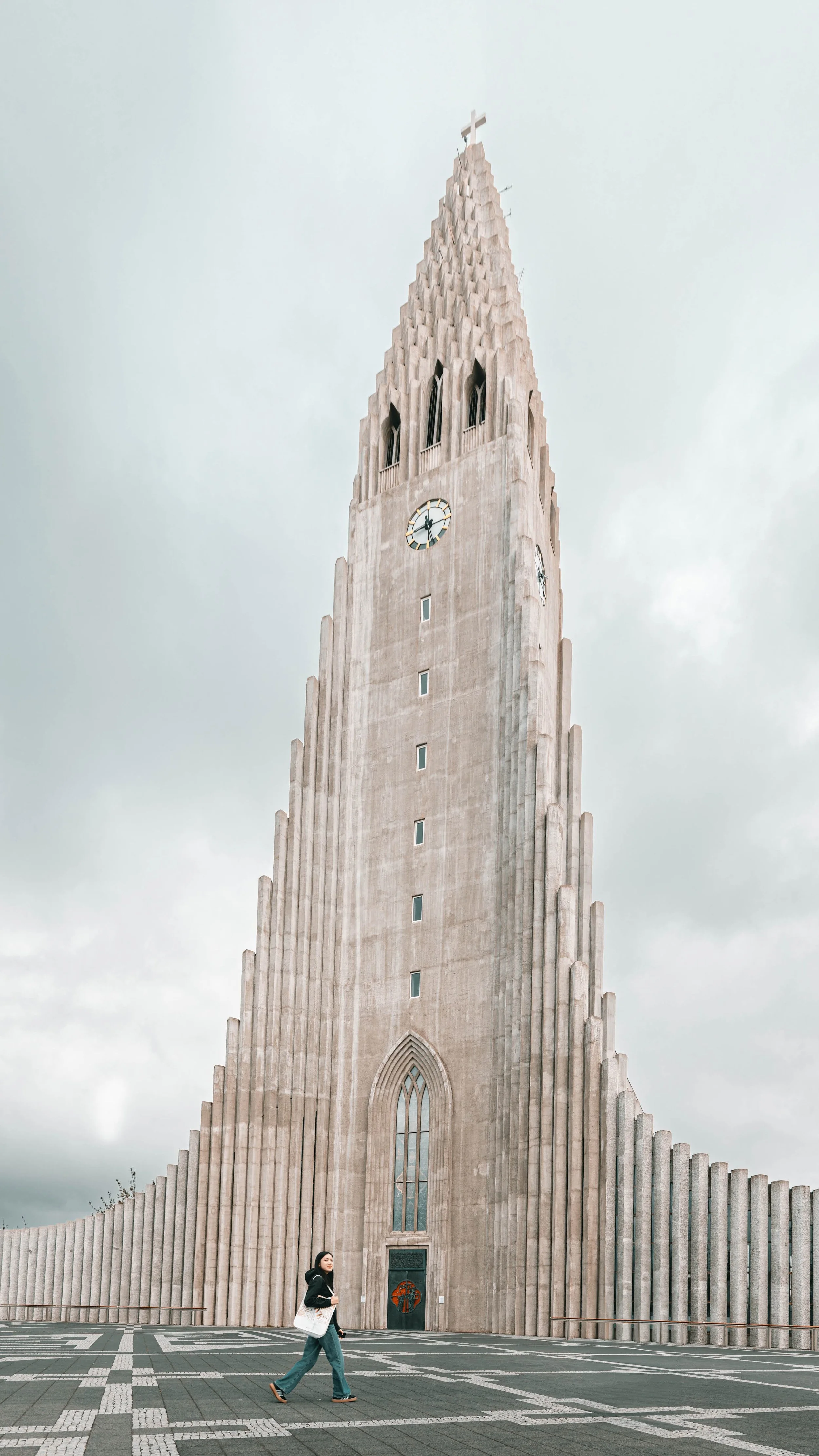 A tall, modern church tower in Reykjavik, Iceland, with a pointed top and cross on it, and a woman walking on the sidewalk in front of it.