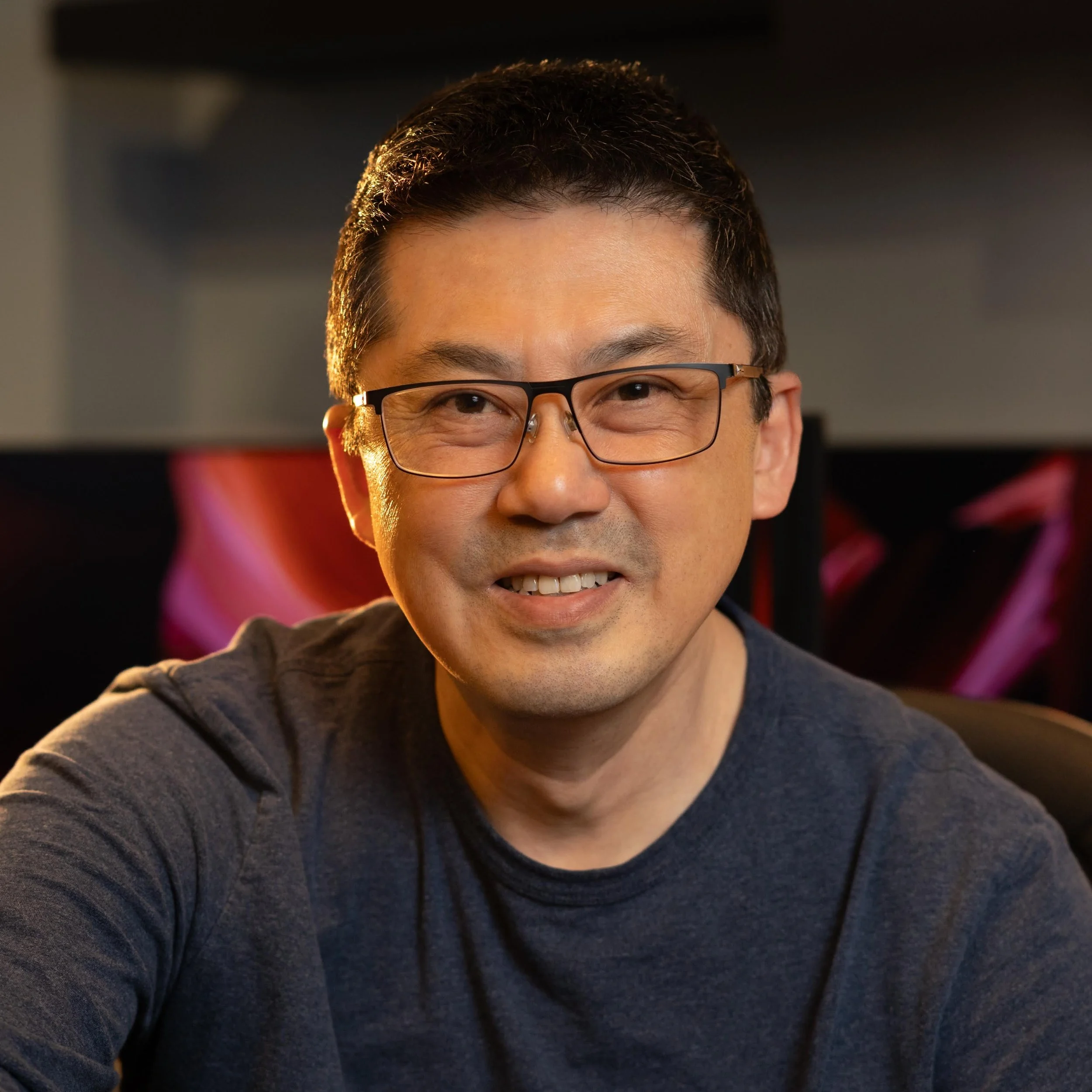 A man with short dark hair, glasses, and a friendly smile, wearing a dark grey shirt, sitting in front of a computer with a colorful monitor.
