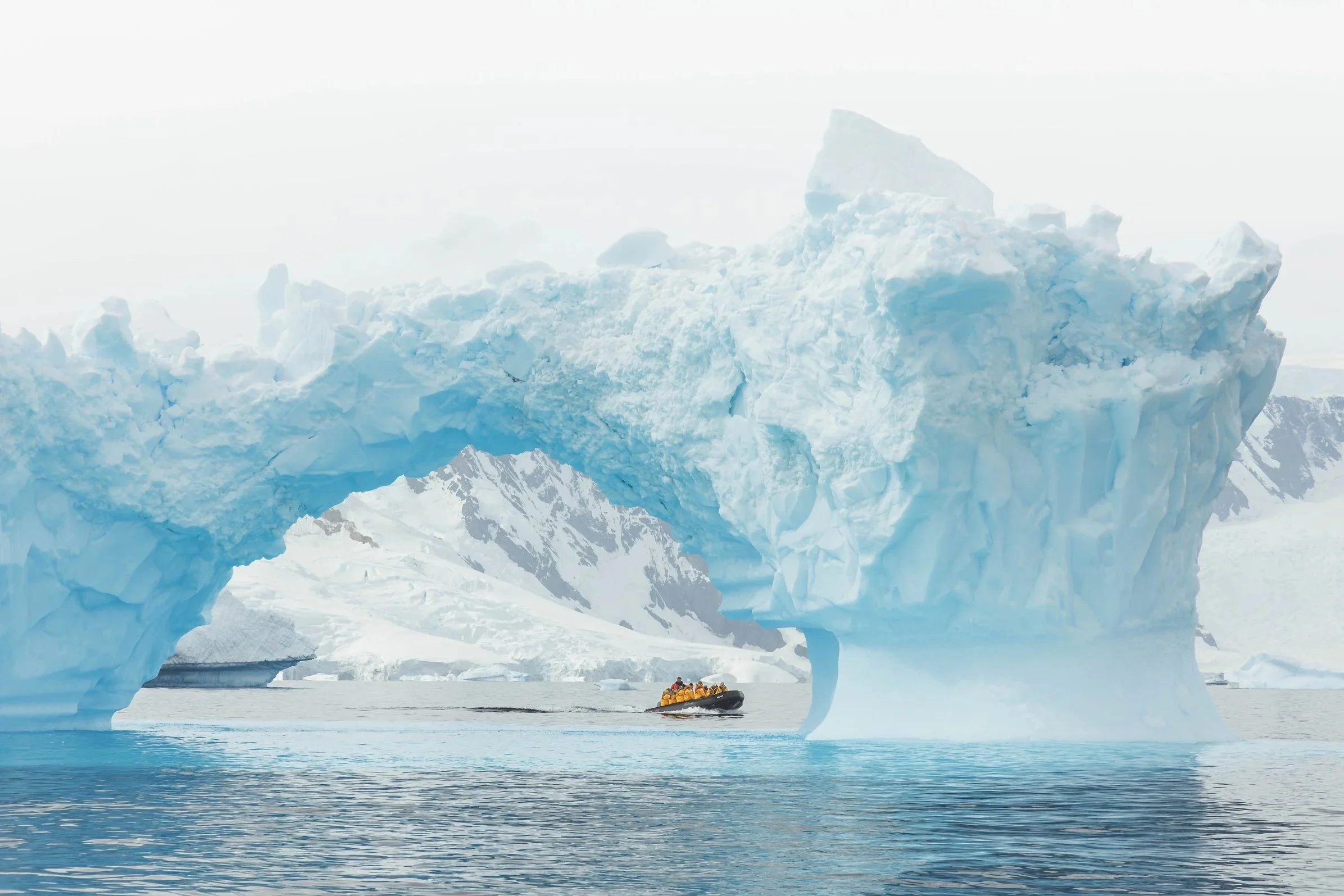 Zodiac full of passengers in yellow jackets passing through the opening of a large blue iceberg arch, Antarctica