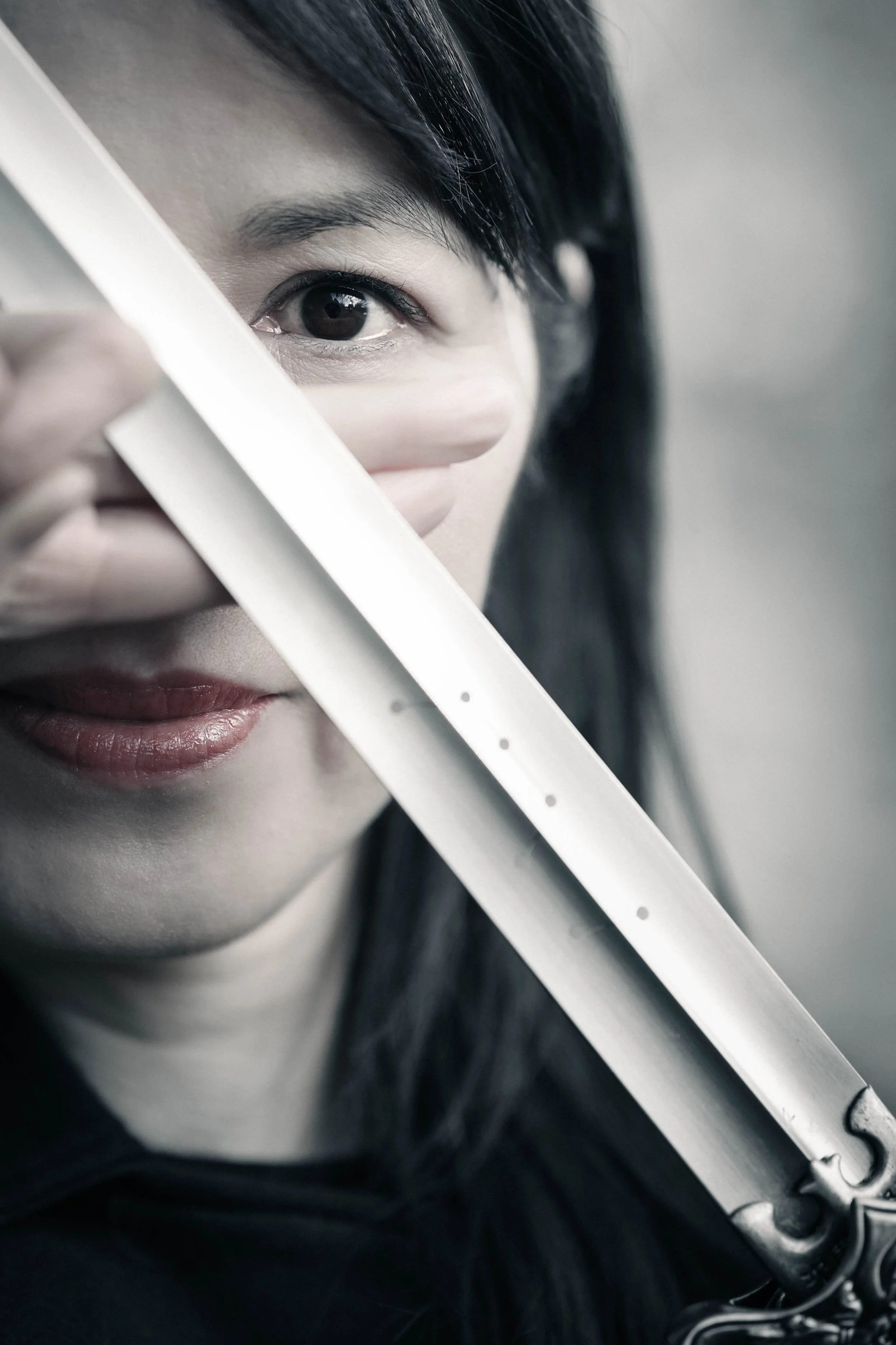 Close-up of a beautiful woman's face with only one eye visible, holding a tai-chi sword diagonally across her face.