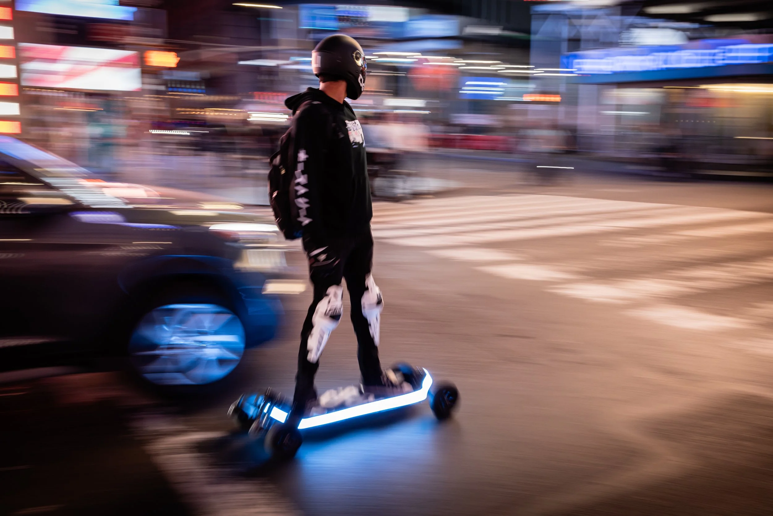 Person riding an electric scooter on a city street at night, wearing a black helmet and dark clothing, with blurred lights and buildings in the background.