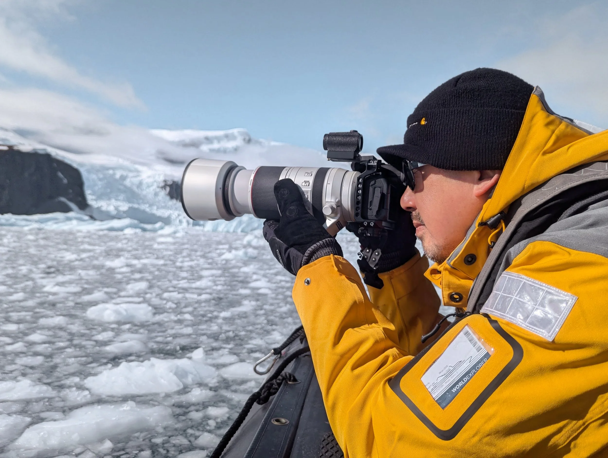 Photographer in a yellow jacket shooting from a Zodiac with a Canon camera, long lens and red dot sight, floating ice and glacier behind