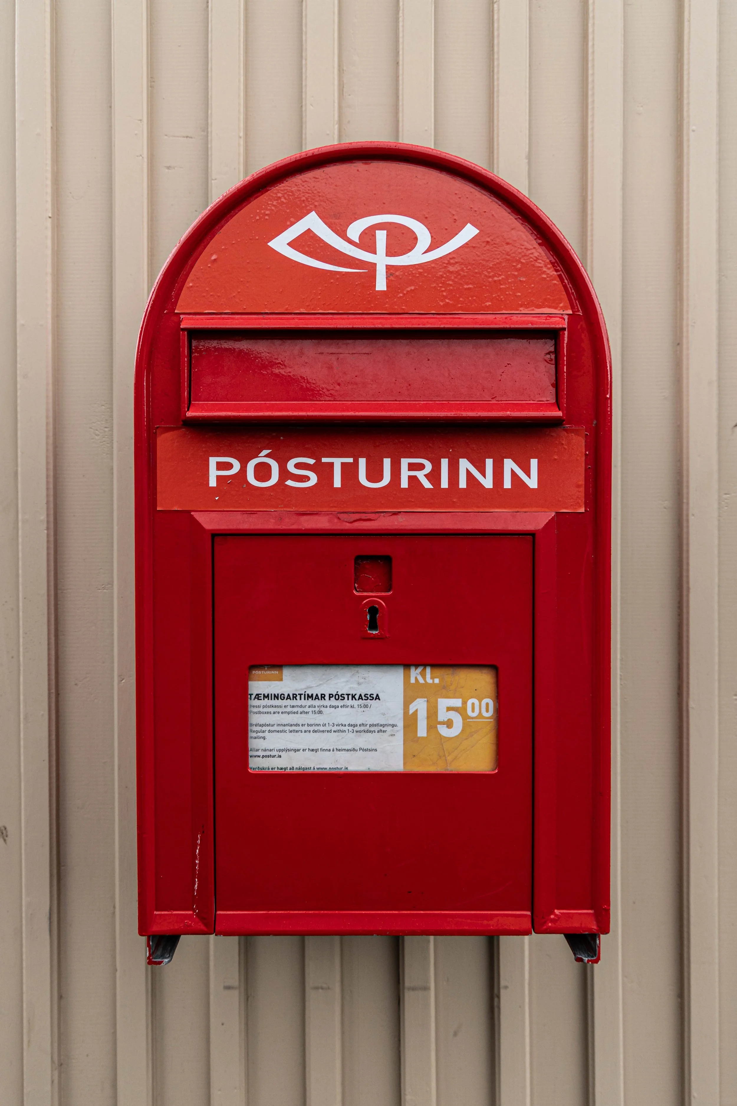 Red Icelandic mailbox mounted on a beige metal wall, with a white symbol on top and the word 'POSTURINN' printed on it, and a sticker showing the price of 15.00.