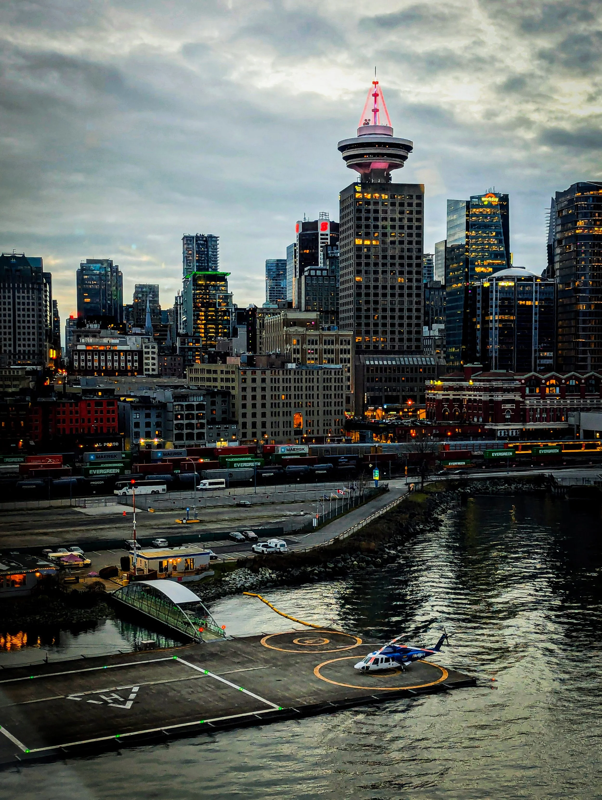 Vancouver downtown skyline during dusk with a Helijet helicopter on a helipad by the water, high-rise buildings with lights on, and a prominent tower with a pink pointed top.