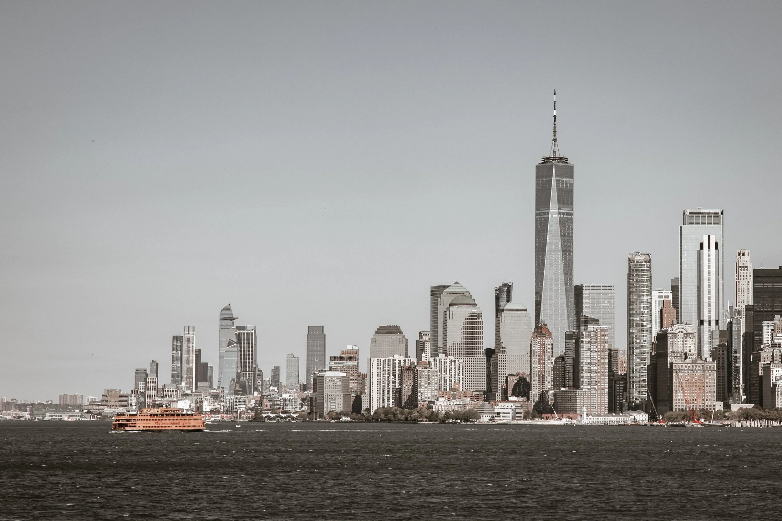 View of New York City skyline with tall skyscrapers, including the One World Trade Center, seen across a body of water with a Staten Island ferry in the foreground.