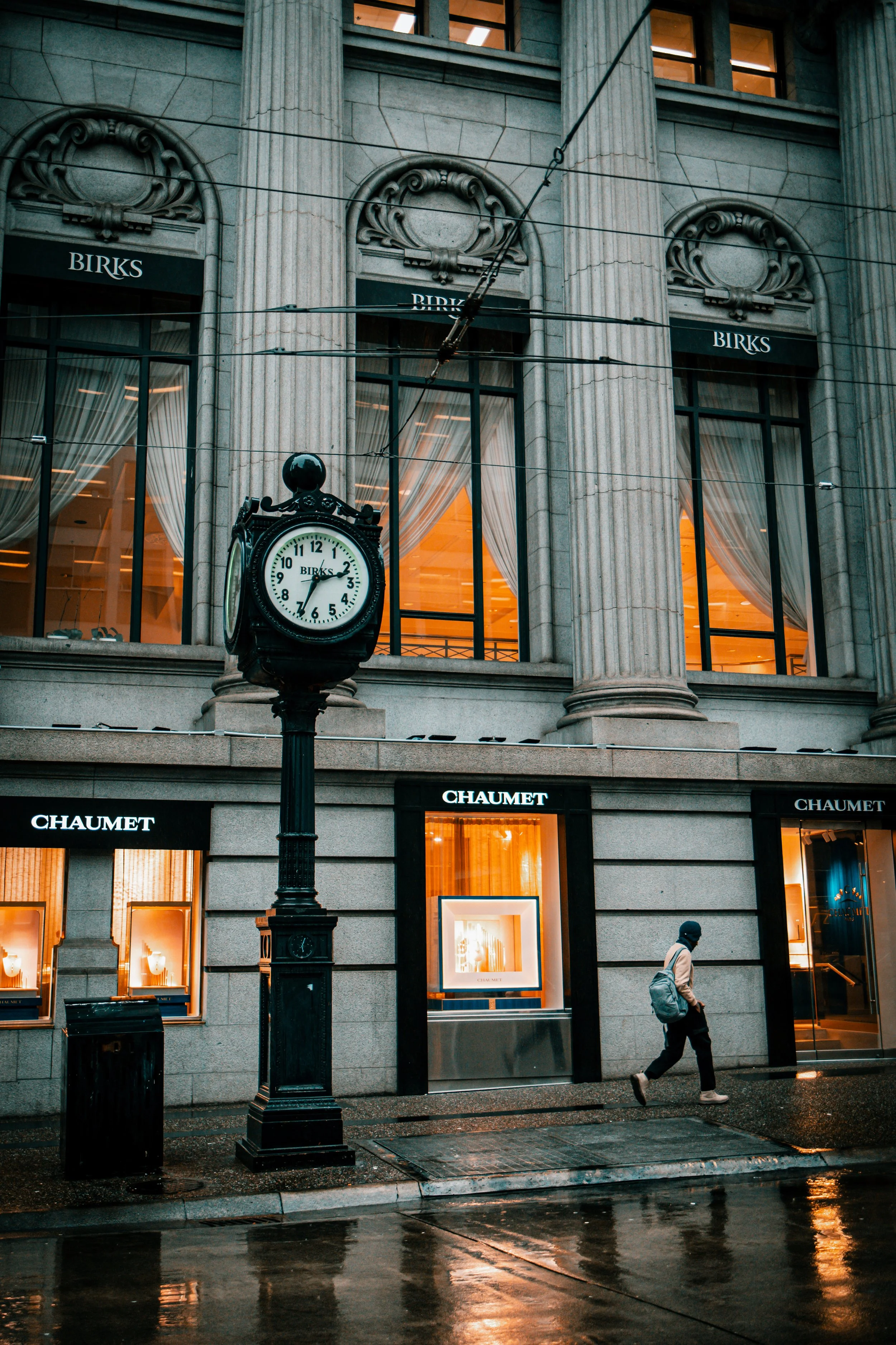 Street scene featuring a person walking on a wet sidewalk, a vintage clock on a pole, and a building with large windows and columns, illuminated with warm lighting; storefront signs reading Chaumet and Birks.