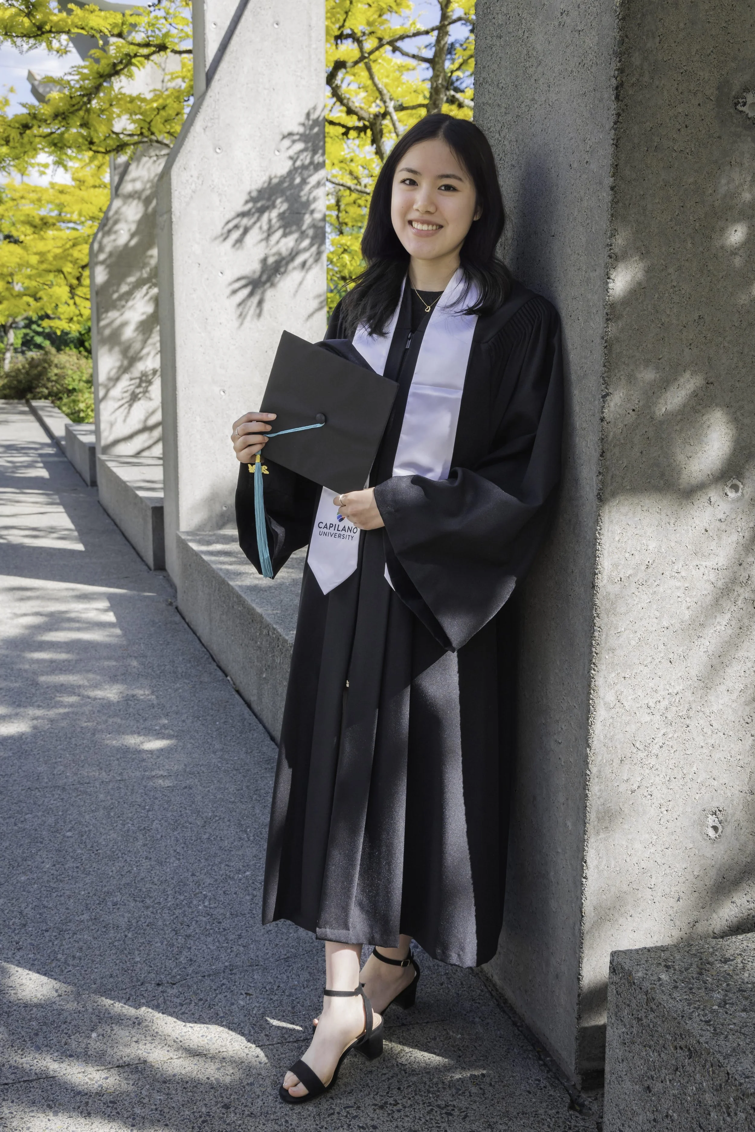 A young woman in a black graduation gown and cap holding a diploma standing outdoors near a concrete wall, with yellow autumn leaves in the background.