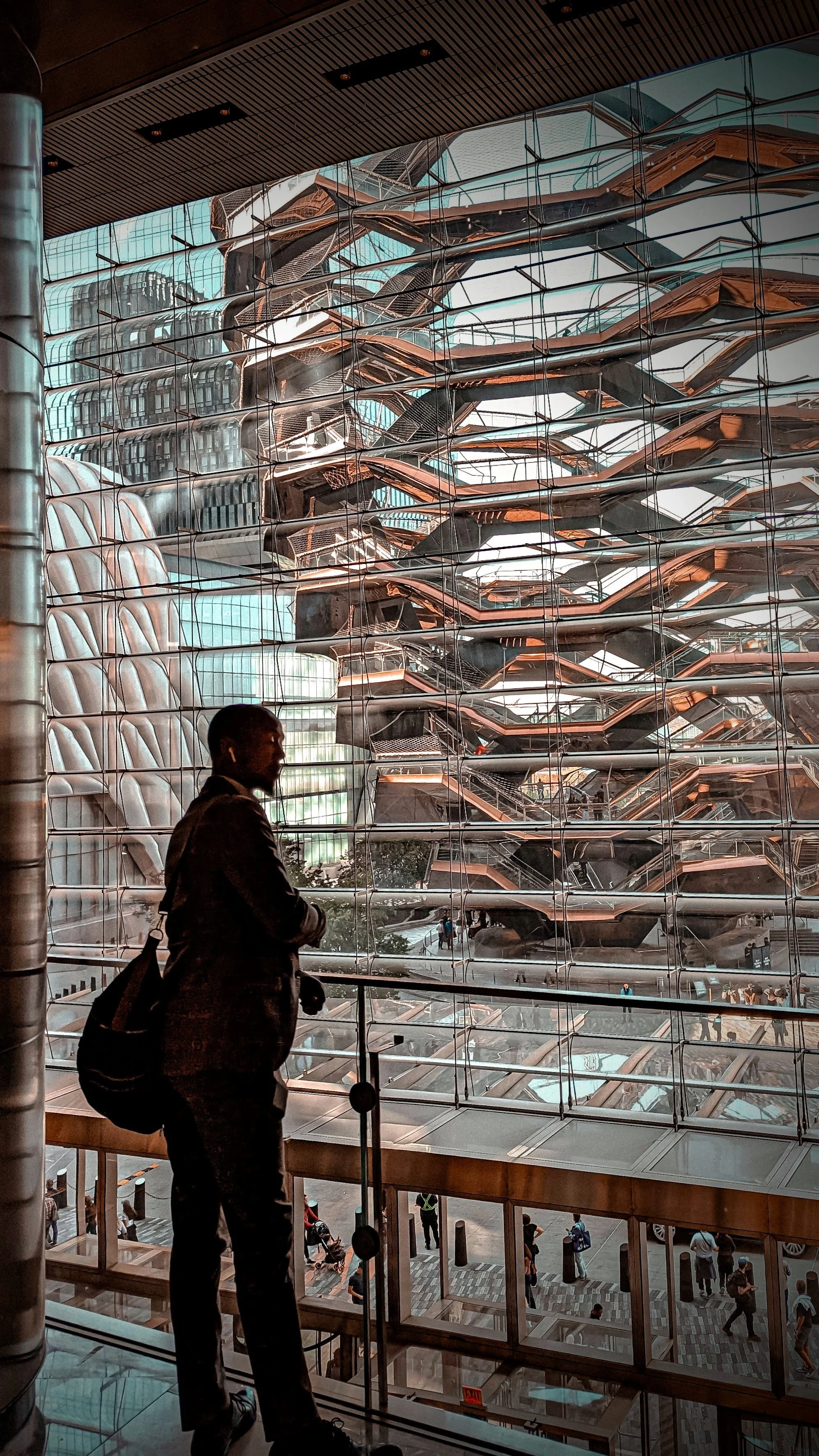 A well-dressed black person standing indoors, looking out a large glass window at the Vessel in New York's Hudson Yards neighborhood, featuring multiple staircases or walkways and a reflective surface.