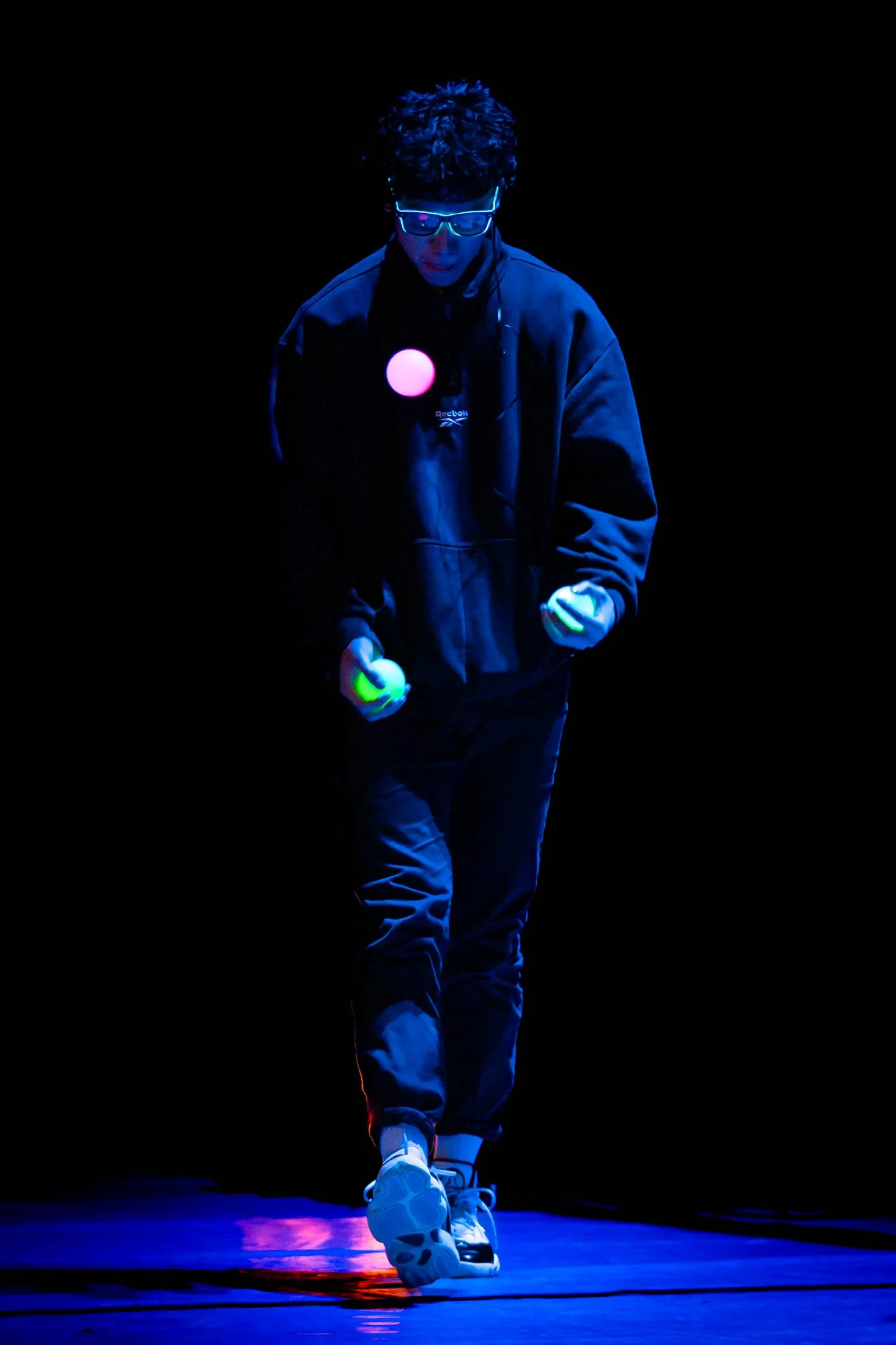 A person with lit neon glasses and curly hair juggling glow-in-the-dark balls on a dark stage, illuminated by ultraviolet light.