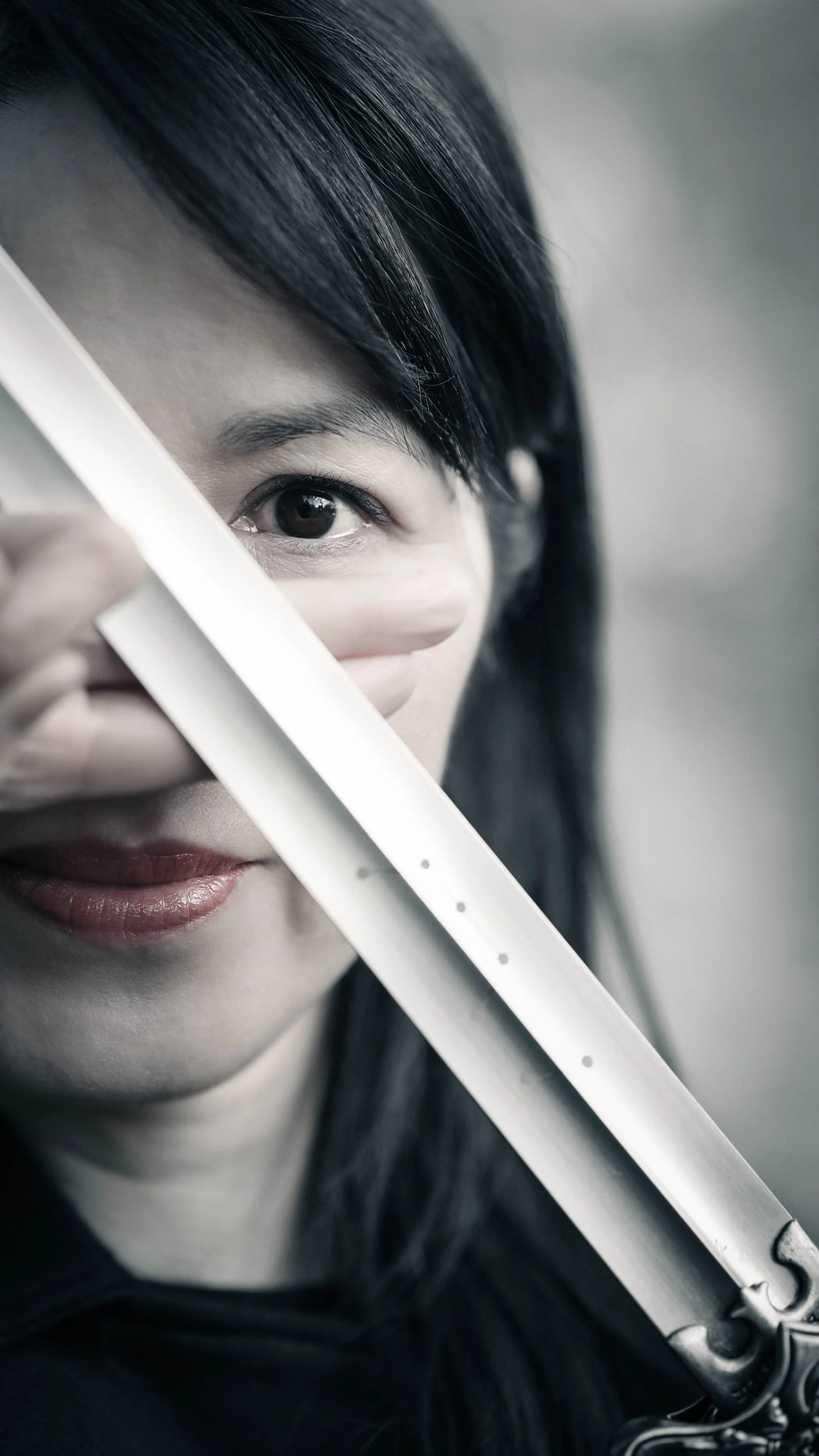 A young woman with dark hair holding a tai-chi sword close to her face, partially covering her mouth, with part of her face and eye visible.