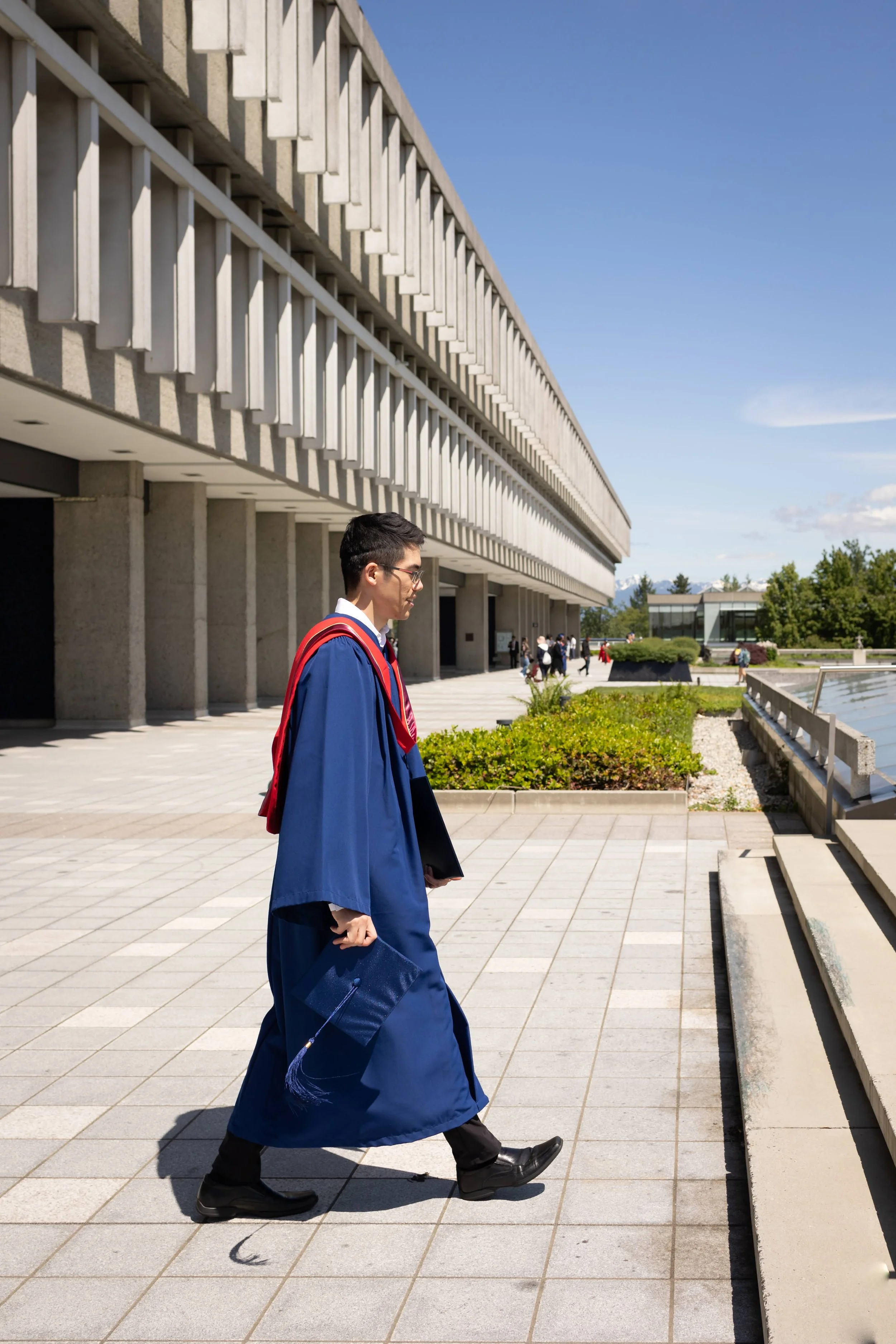 A young man in a blue graduation gown and cap walking outside near a modern building and water feature on a sunny day.