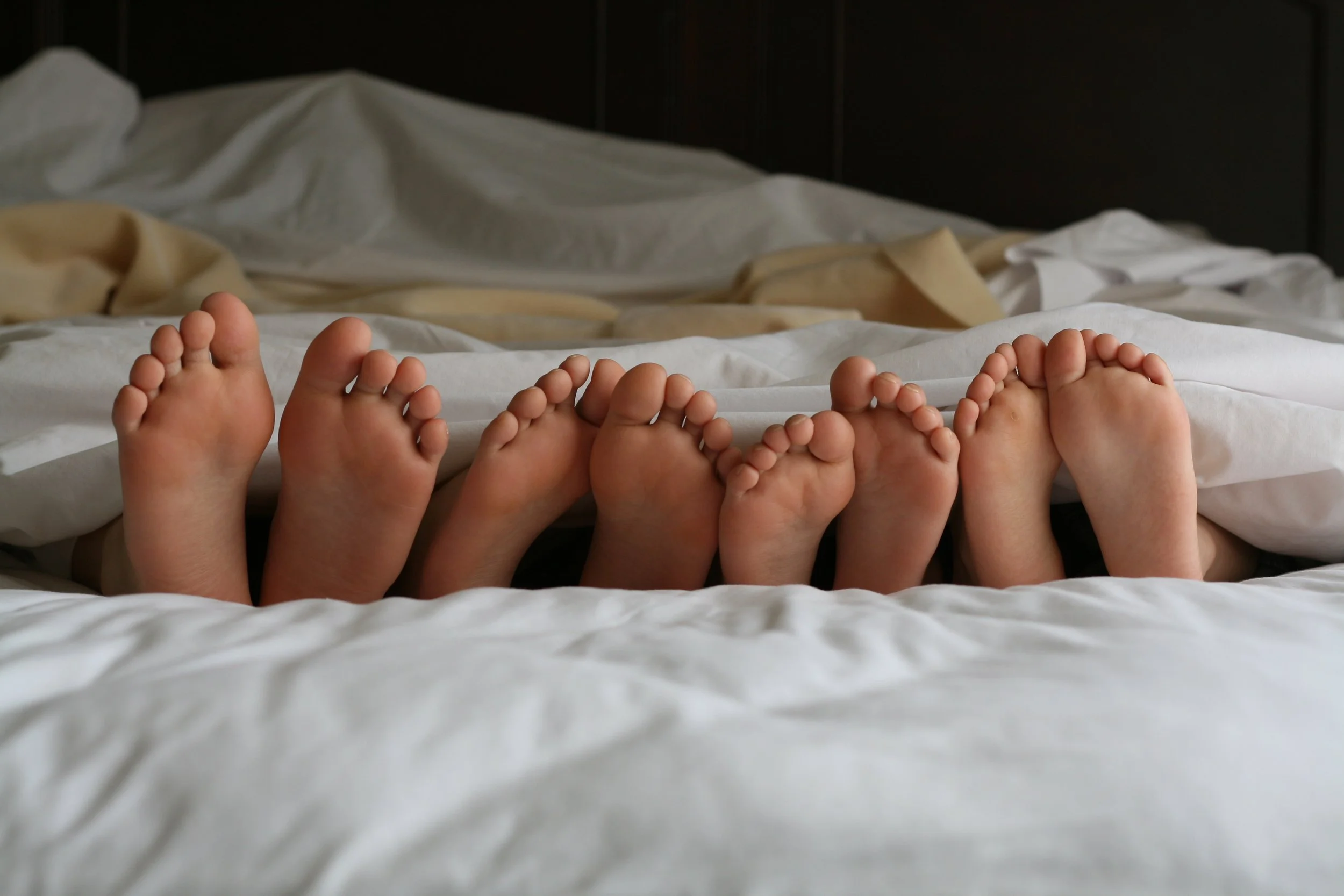 Multiple pairs of bare kid's feet sticking out from under a bed sheet on a bed with white bedding.