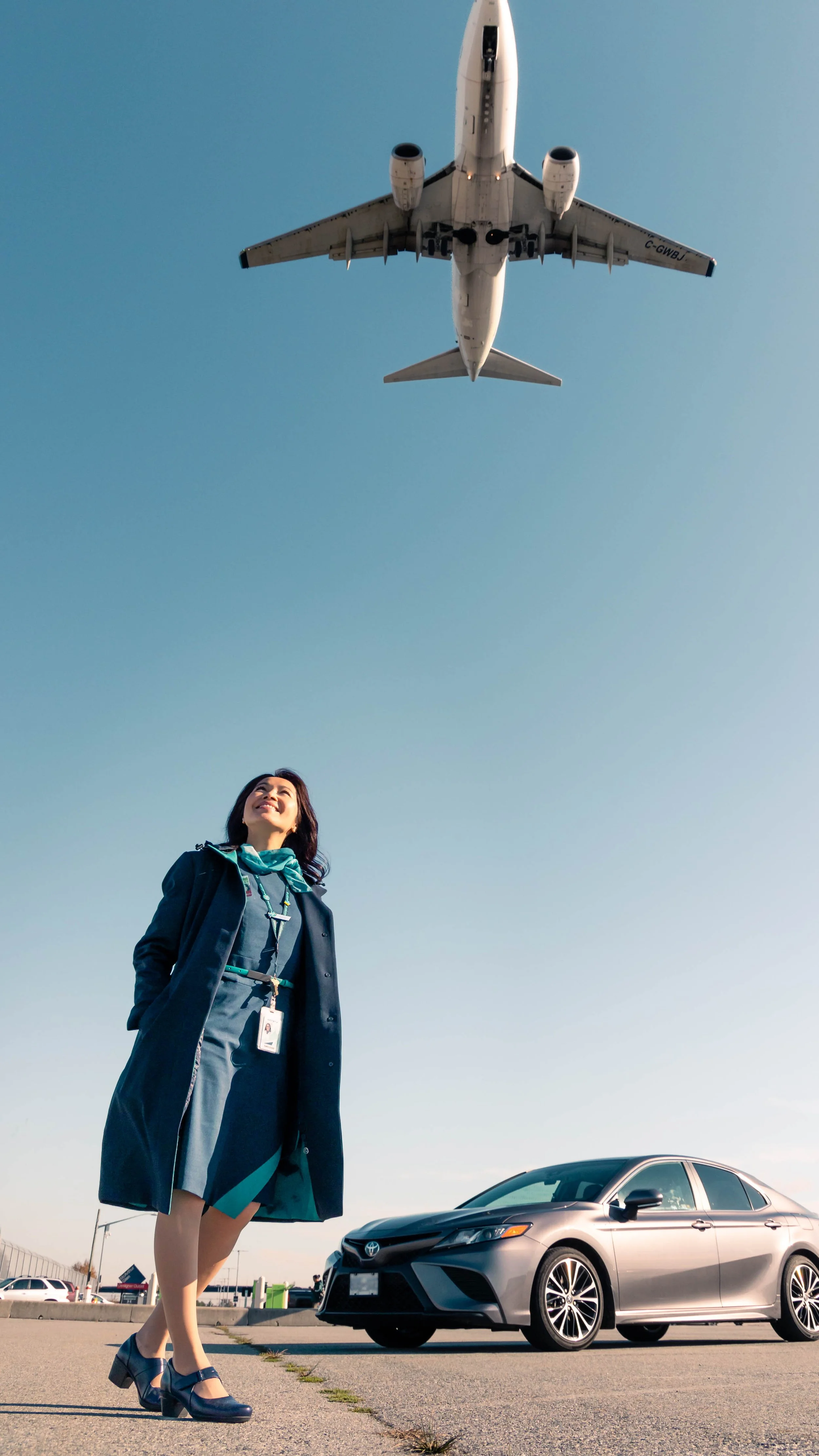 A woman standing on an airport tarmac looking up at a plane flying overhead, with a parked silver car nearby.