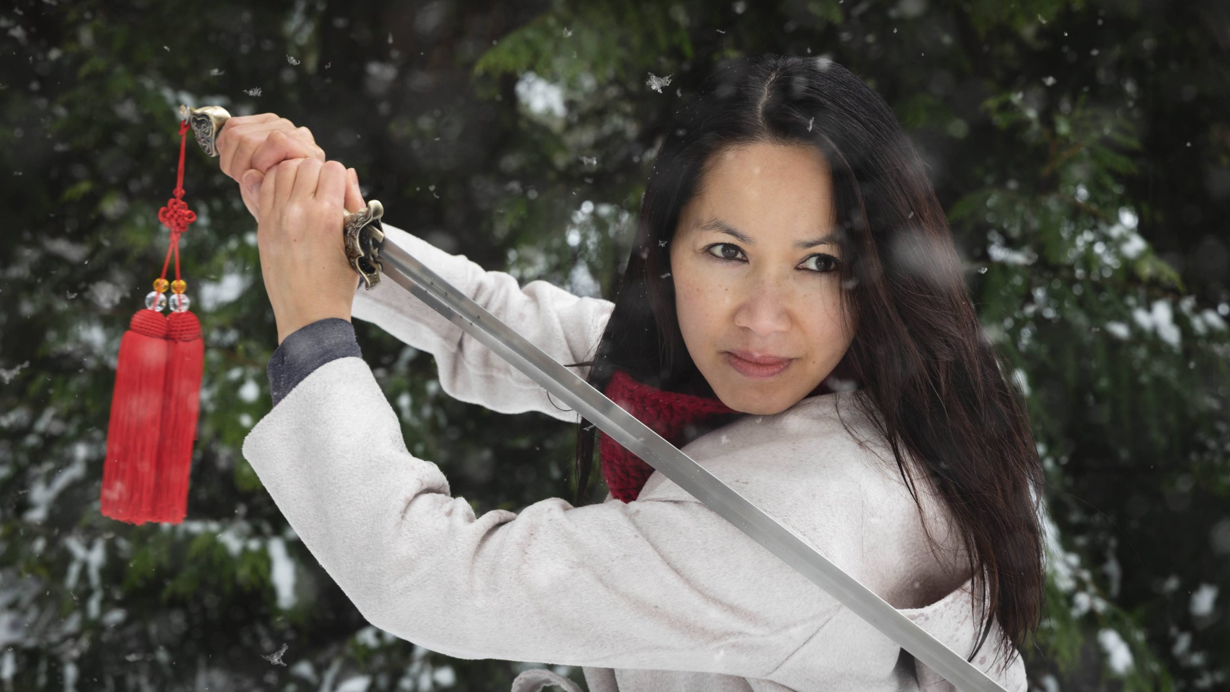Woman with long dark hair in a white coat holding a tai-chi sword outdoors in a snowy forest.