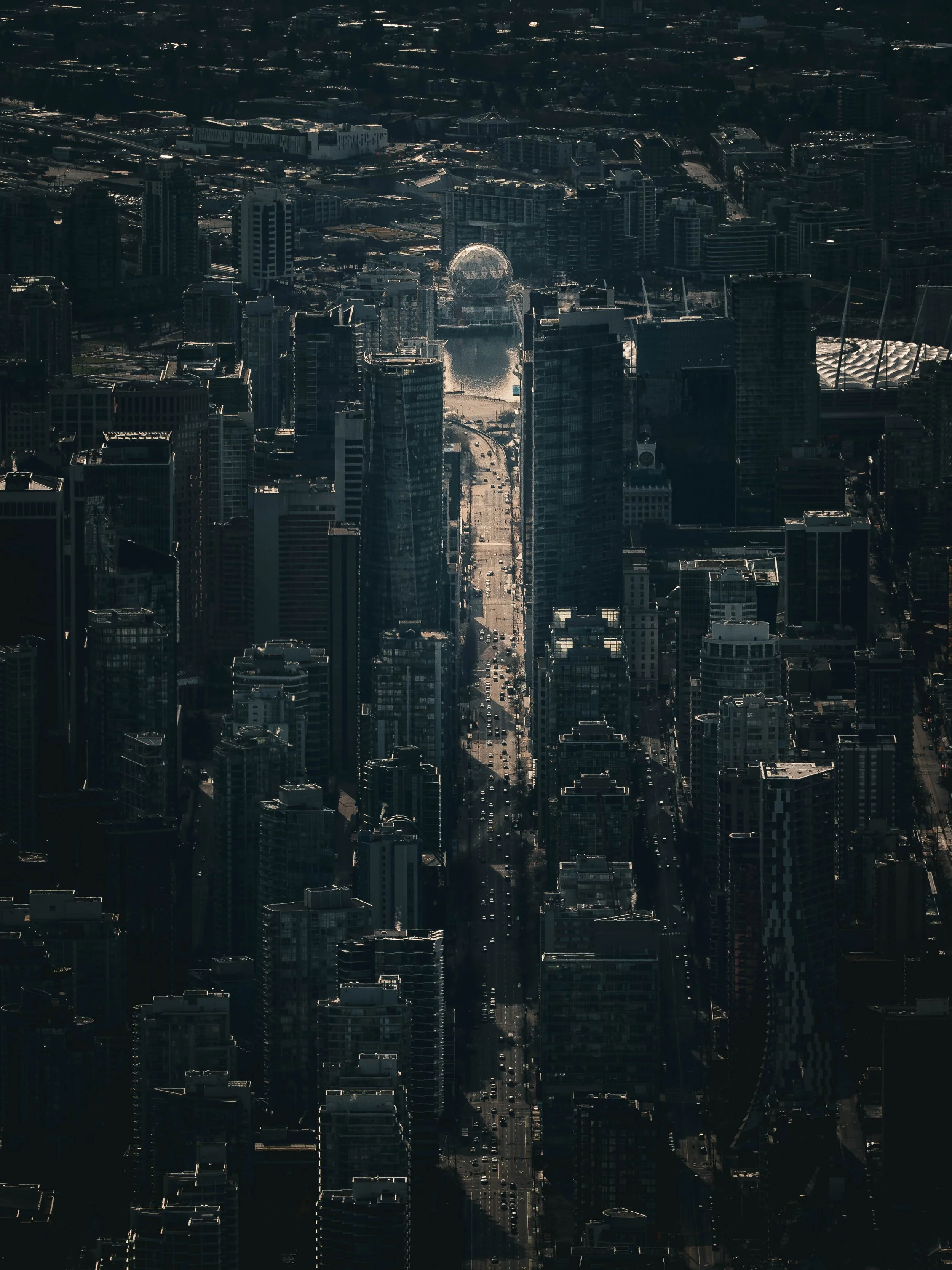 Aerial view of a city with skyscrapers and a body of water reflecting the buildings, with a prominent geodesic dome (Vancouver's Science World) near the water.