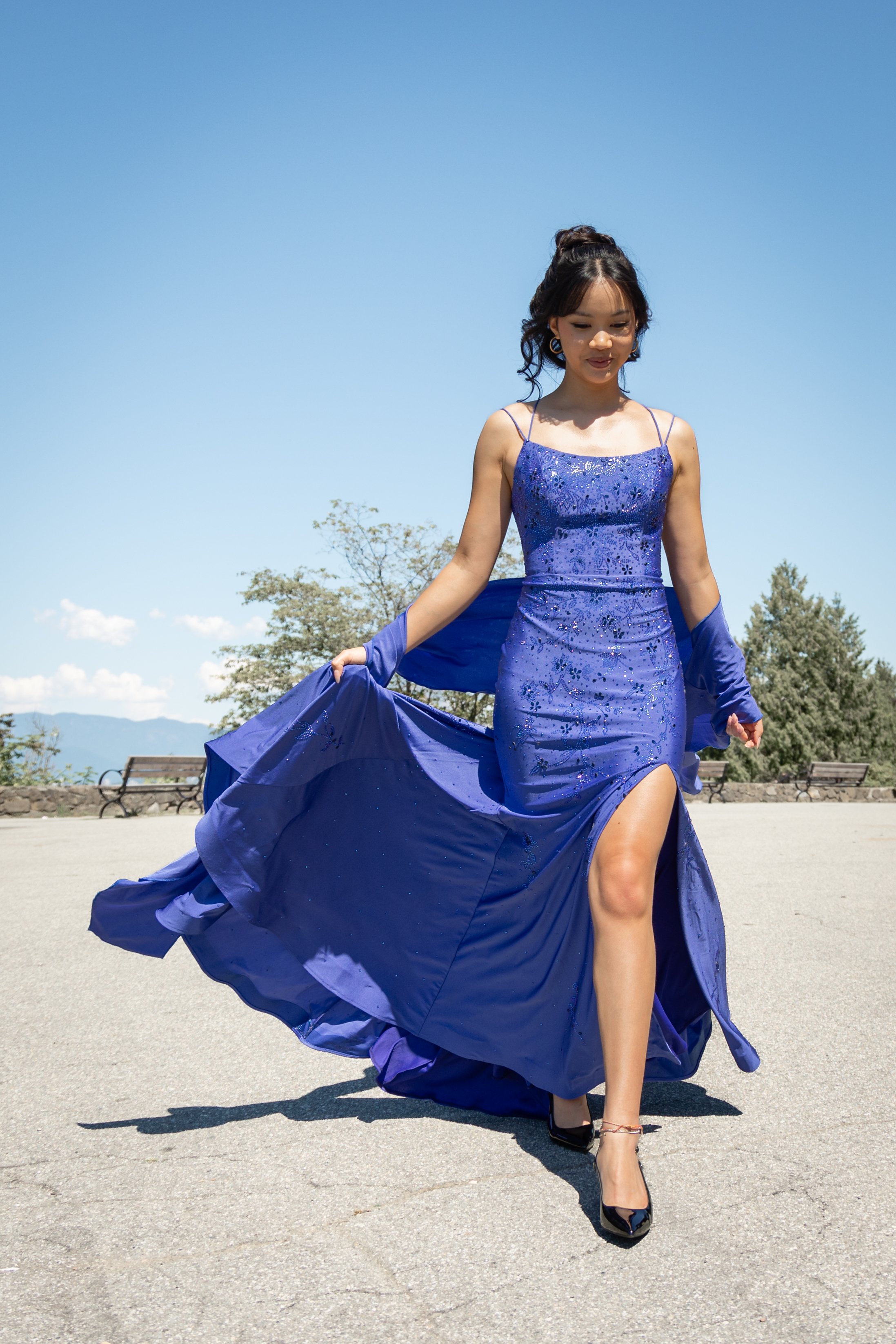A woman in a long, elegant, blue dress with a high slit, walking outdoors on a sunny day.