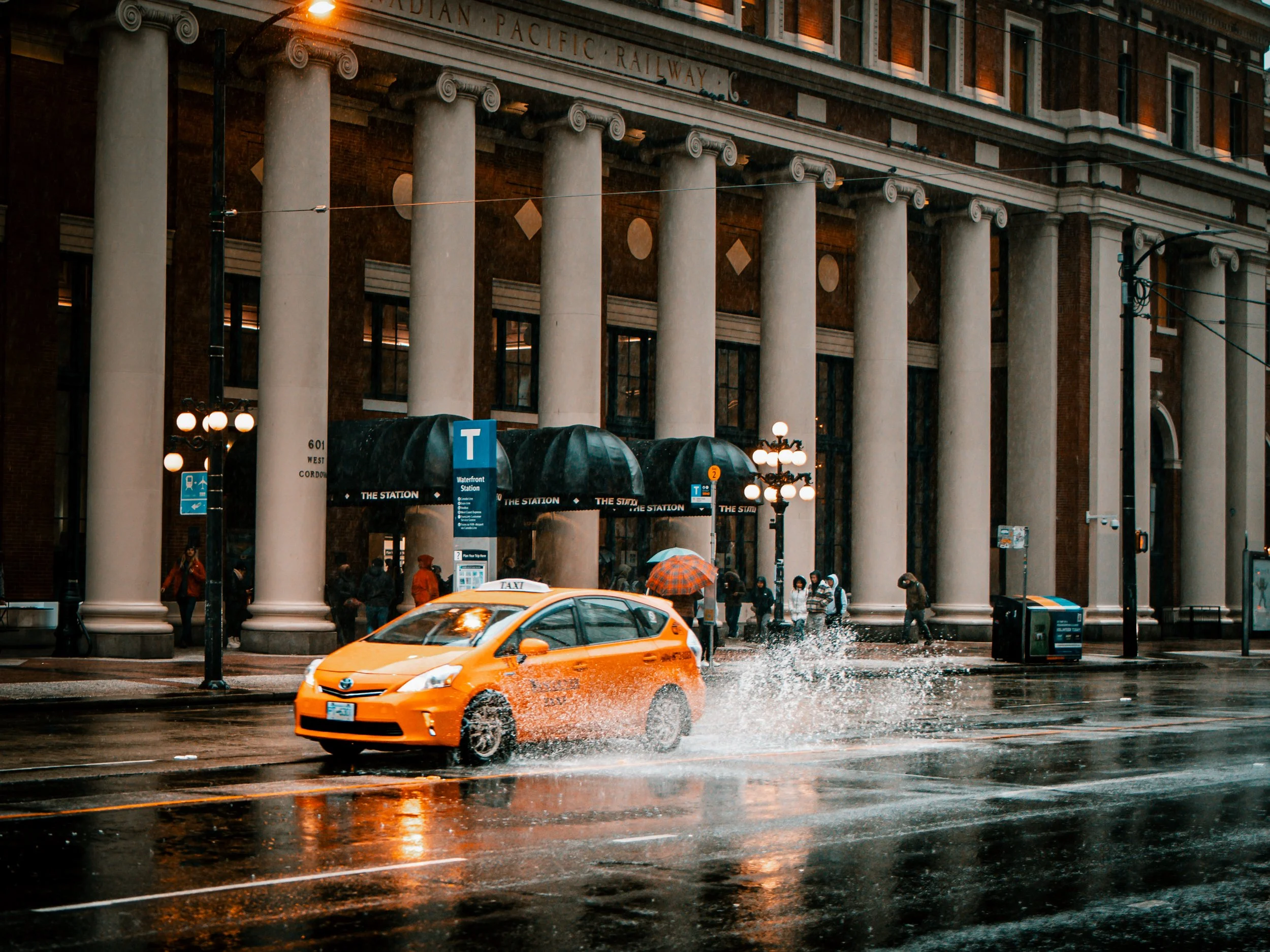 A yellow taxi driving through a wet city street in front of a large building with tall columns and a sign for a water station, with pedestrians under umbrellas during rainy weather at dusk.