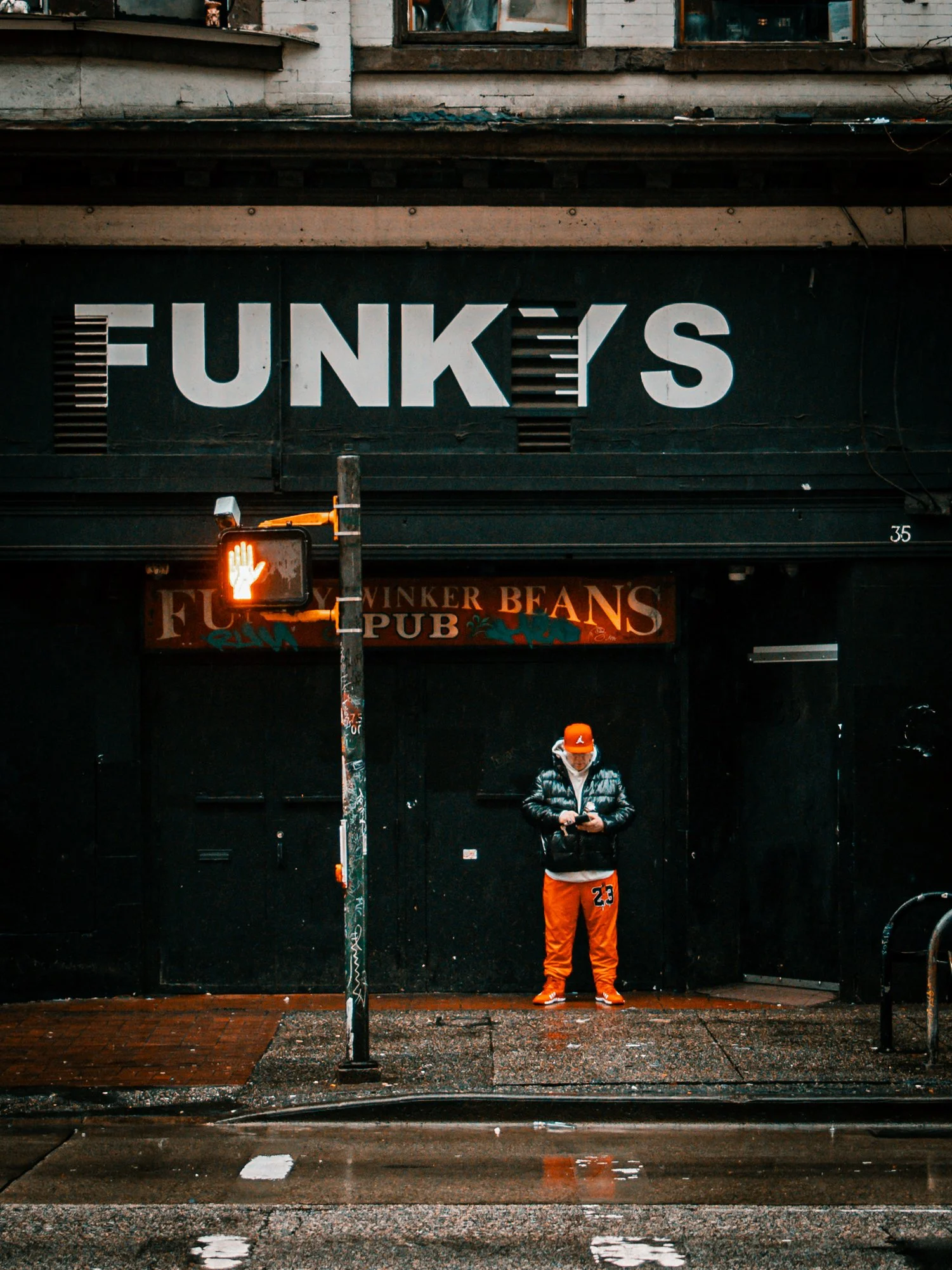 A person standing on a wet sidewalk, wearing an orange cap, black jacket, and orange pants, looking at their phone in front of a dark building with a large sign that says 'FUNKEY'S' and a smaller sign for 'FUNKER BEANS PUB'. There is a red pedestrian