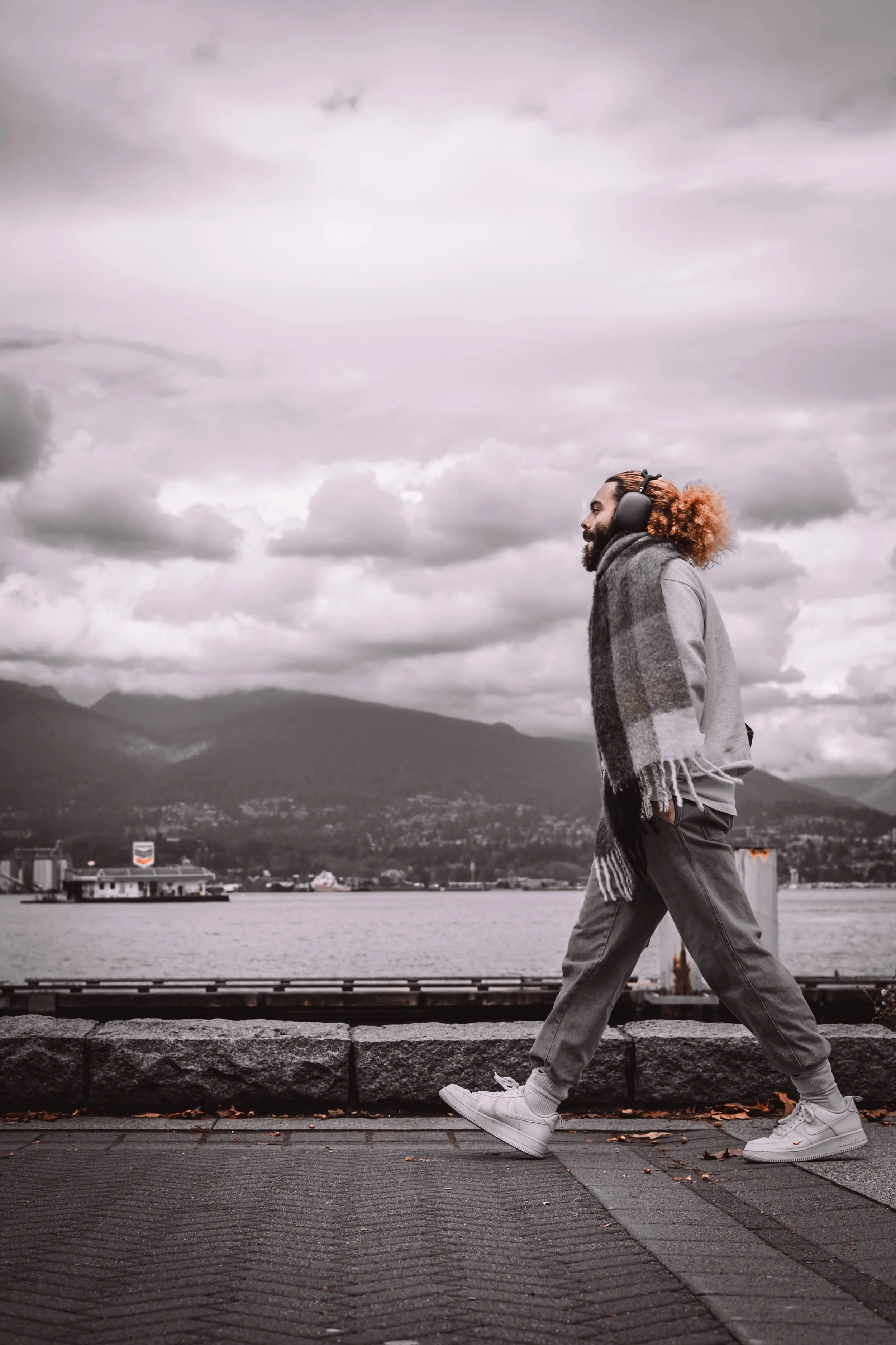 A black man with dreadlocks is walking along a waterfront sidewalk. He is wearing large headphones, a gray hoodie, a plaid scarf, and sneakers. The background features water, mountains, and a cloudy sky.