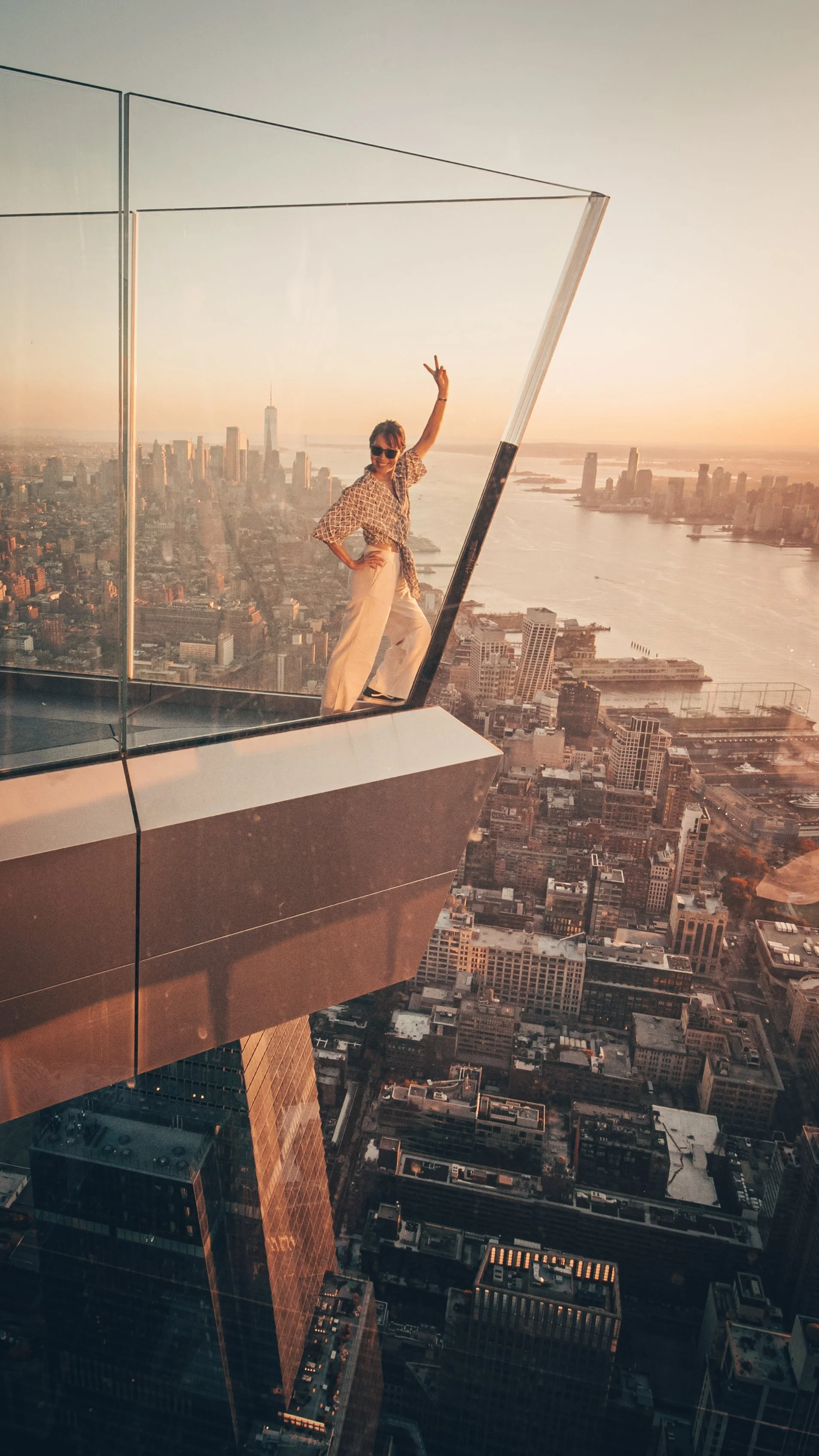 A woman standing on the edge of a glass observation deck of a skyscraper in New York City during sunset, making a disco gesture and peace sign with her arm raised, with the city skyline and river in the background.