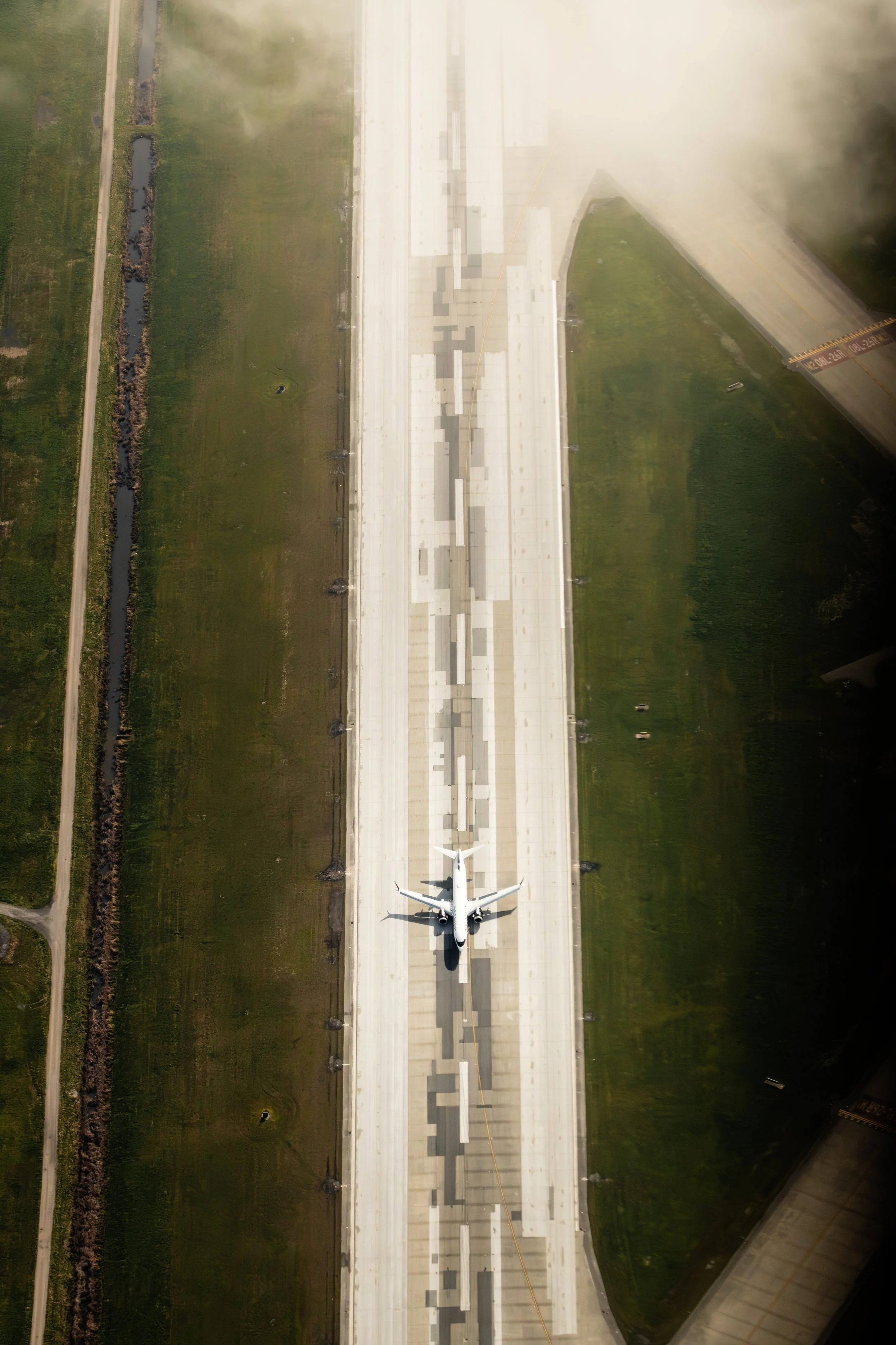 An airplane on a runway seen from above, with green fields on either side and a cloudy sky reflection in the upper right corner.