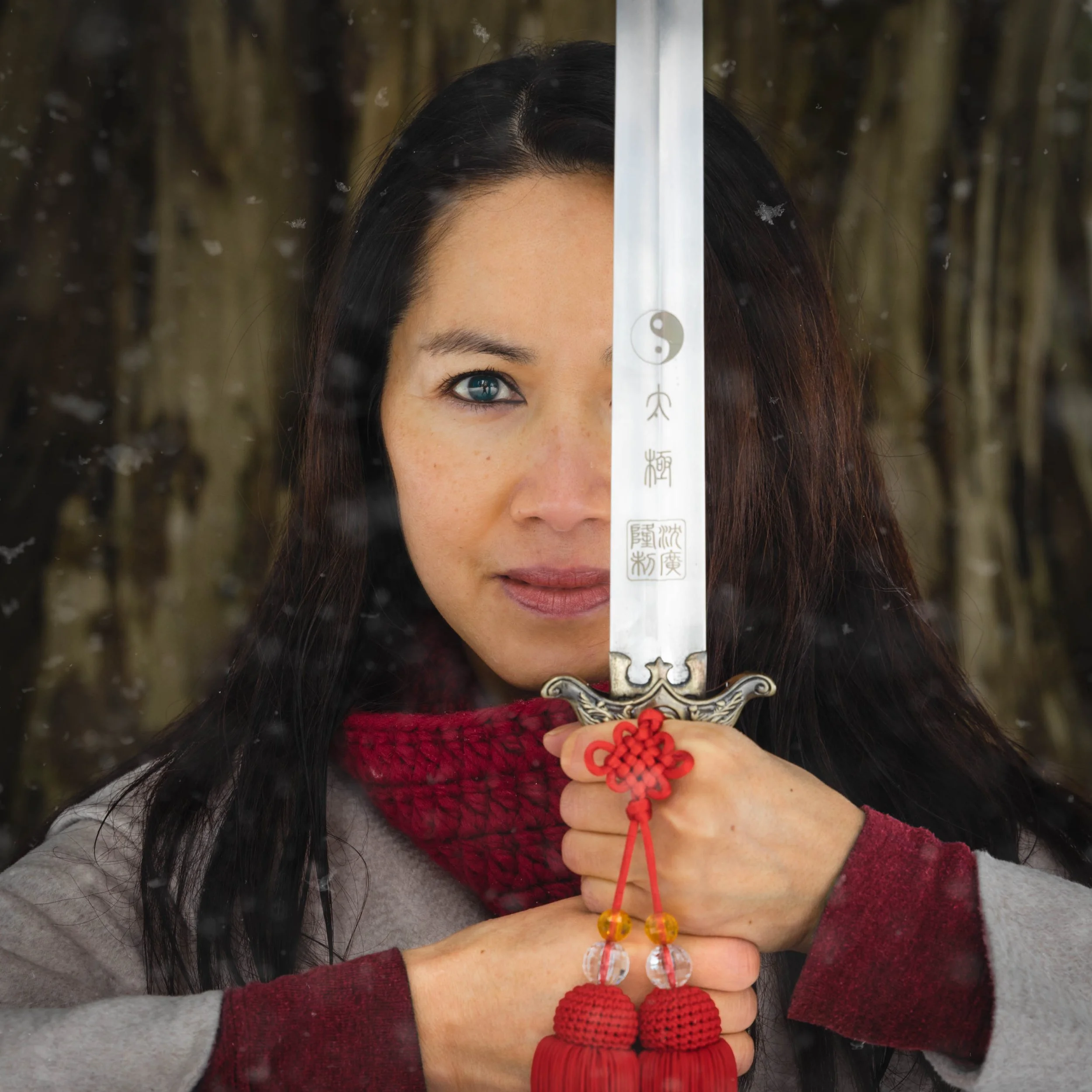 A woman with blue eyes and dark hair holds a tai-chi sword vertically in front of her face, with the blade covering one eye. She wears a red knit scarf, a grey coat, and has a determined expression. The background is blurred with brown bamboo stalks.
