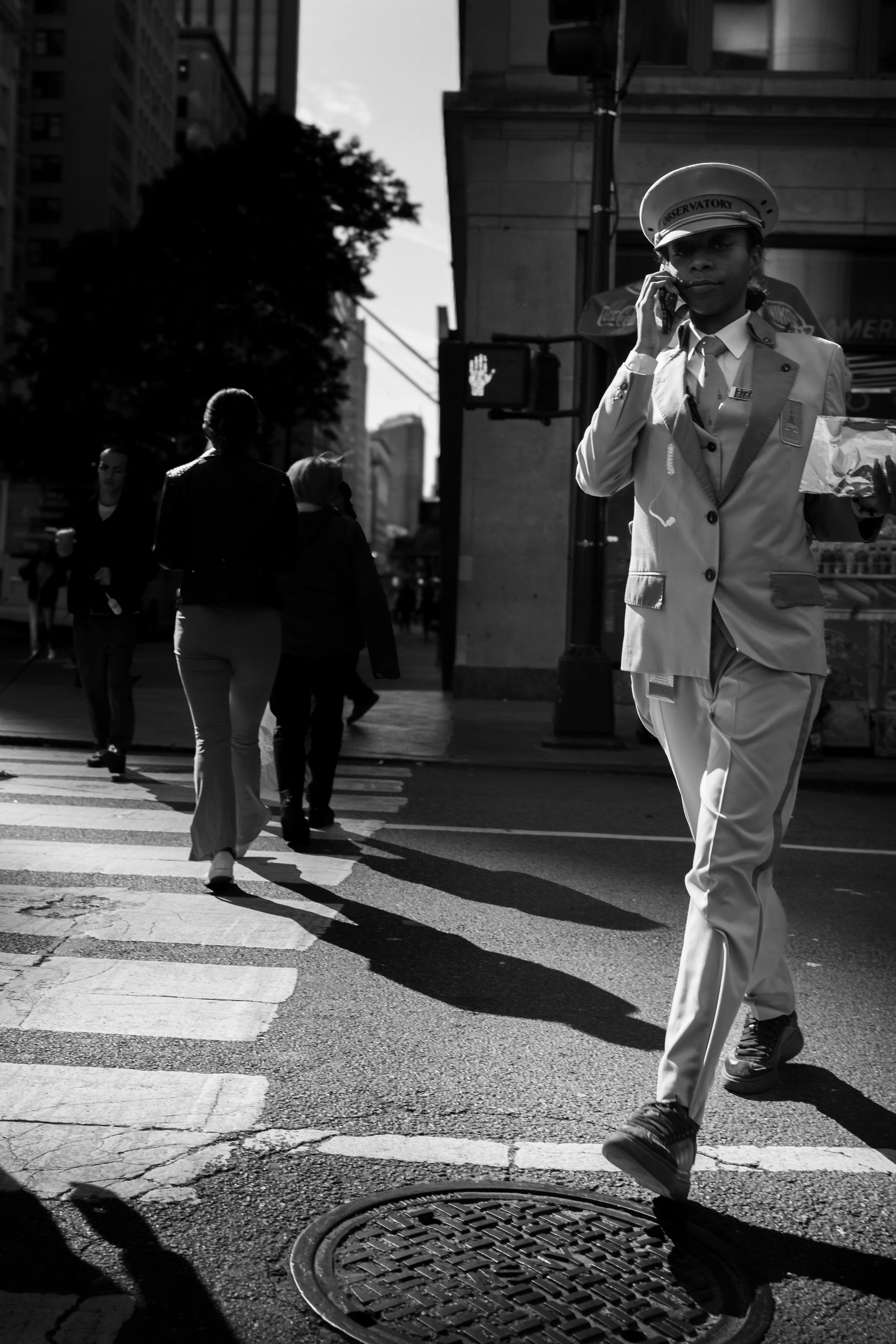 A city street scene in black and white with pedestrians crossing. A woman in a uniform with a peaked cap, talking on a cell phone, is walking toward the camera. Several other people are walking in the opposite direction, and tall buildings are visibl