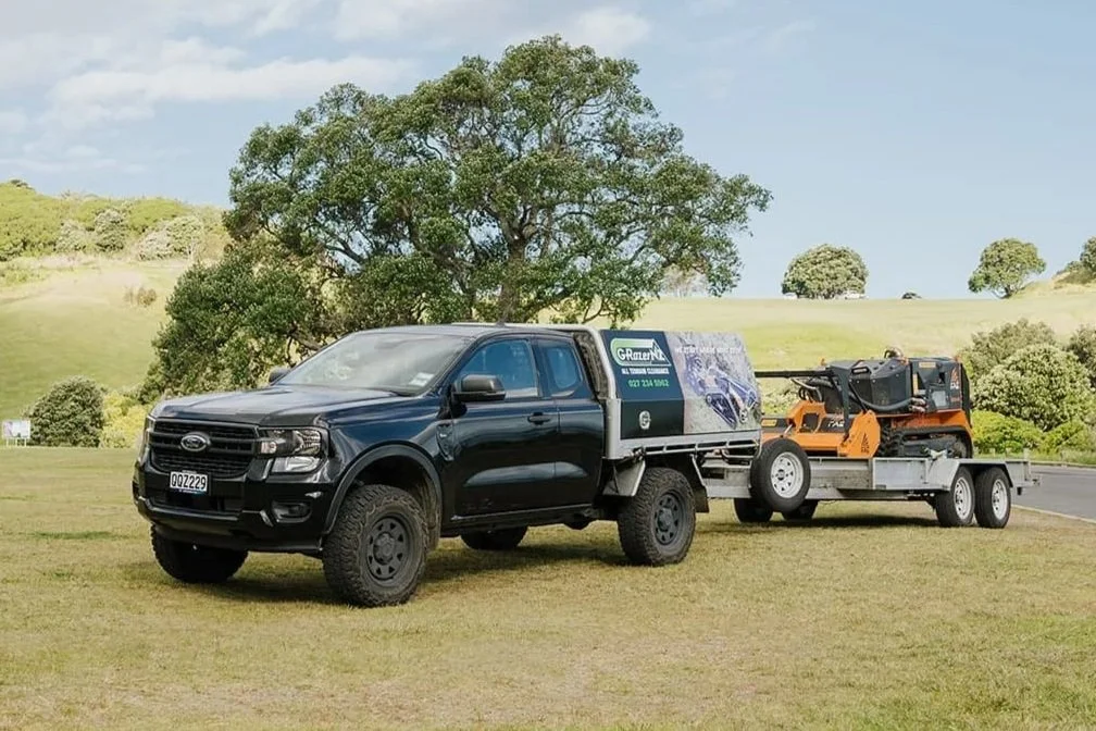 Black pickup truck pulling a trailer carrying a skid steer loader in a grassy field with green hills and trees in the background.