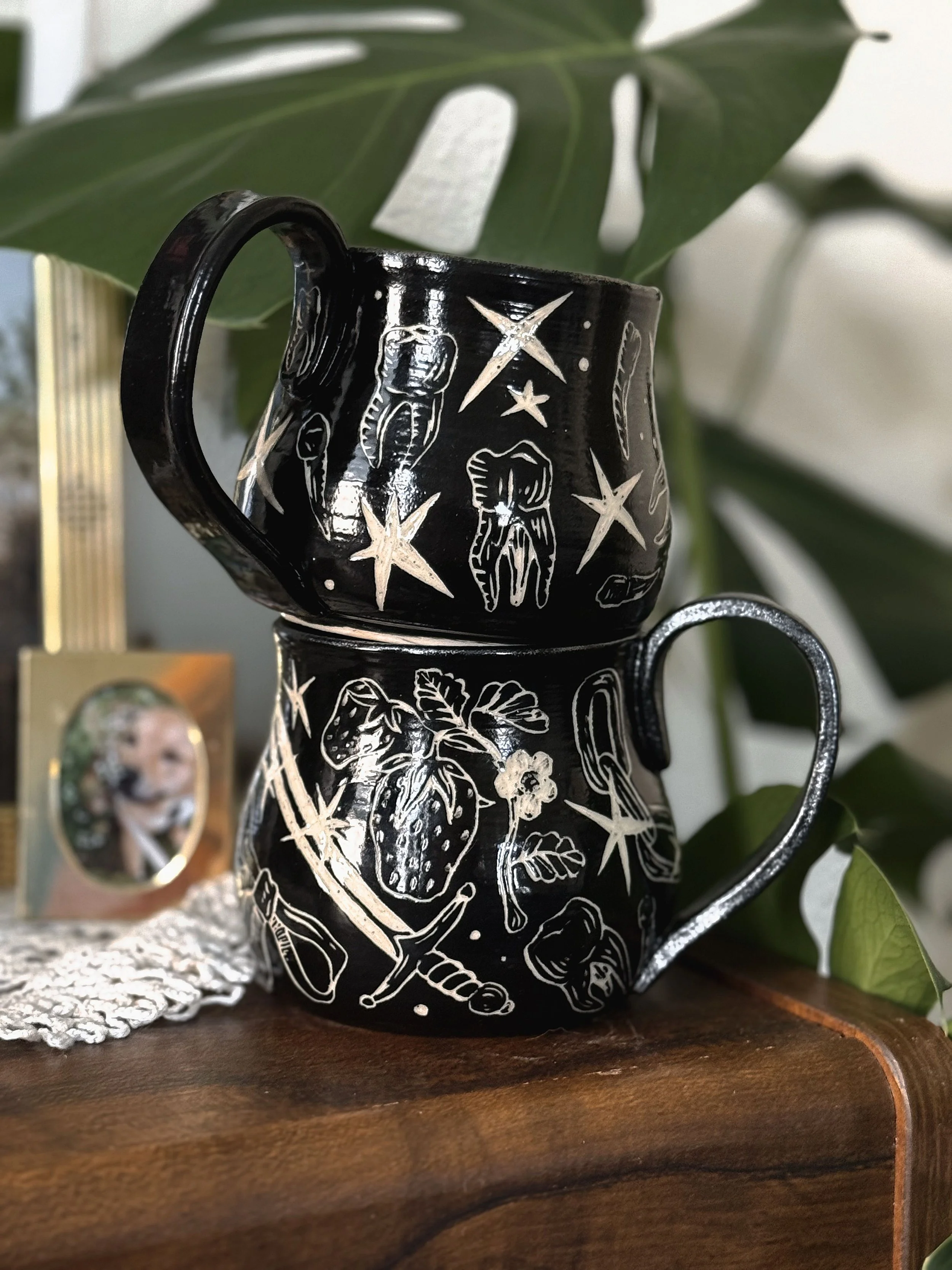 Two black ceramic mugs with various oddity sgraffito carvings.
