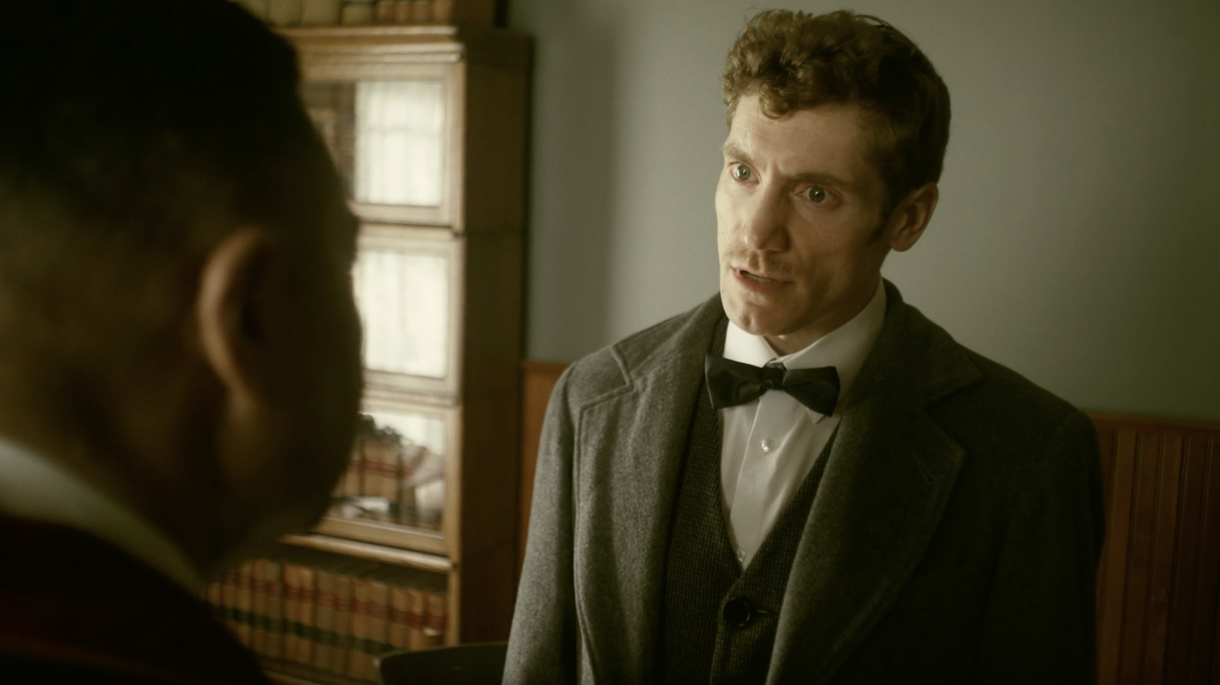 A white man with auburn curly hair wearing a bowtie and black peacoat