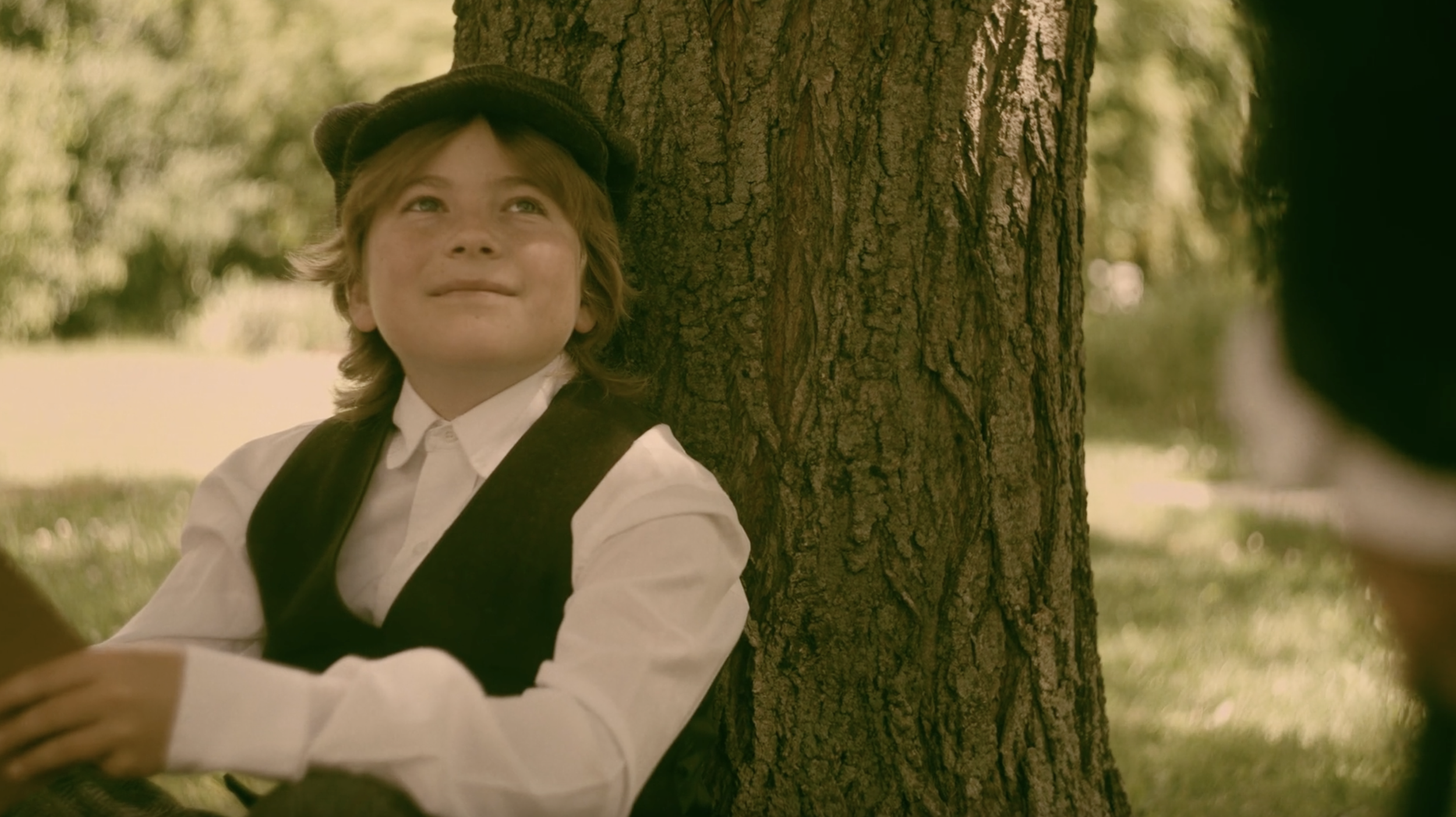 A young boy with shoulder length hair in a newsboy cap and vest sitting under a tree.