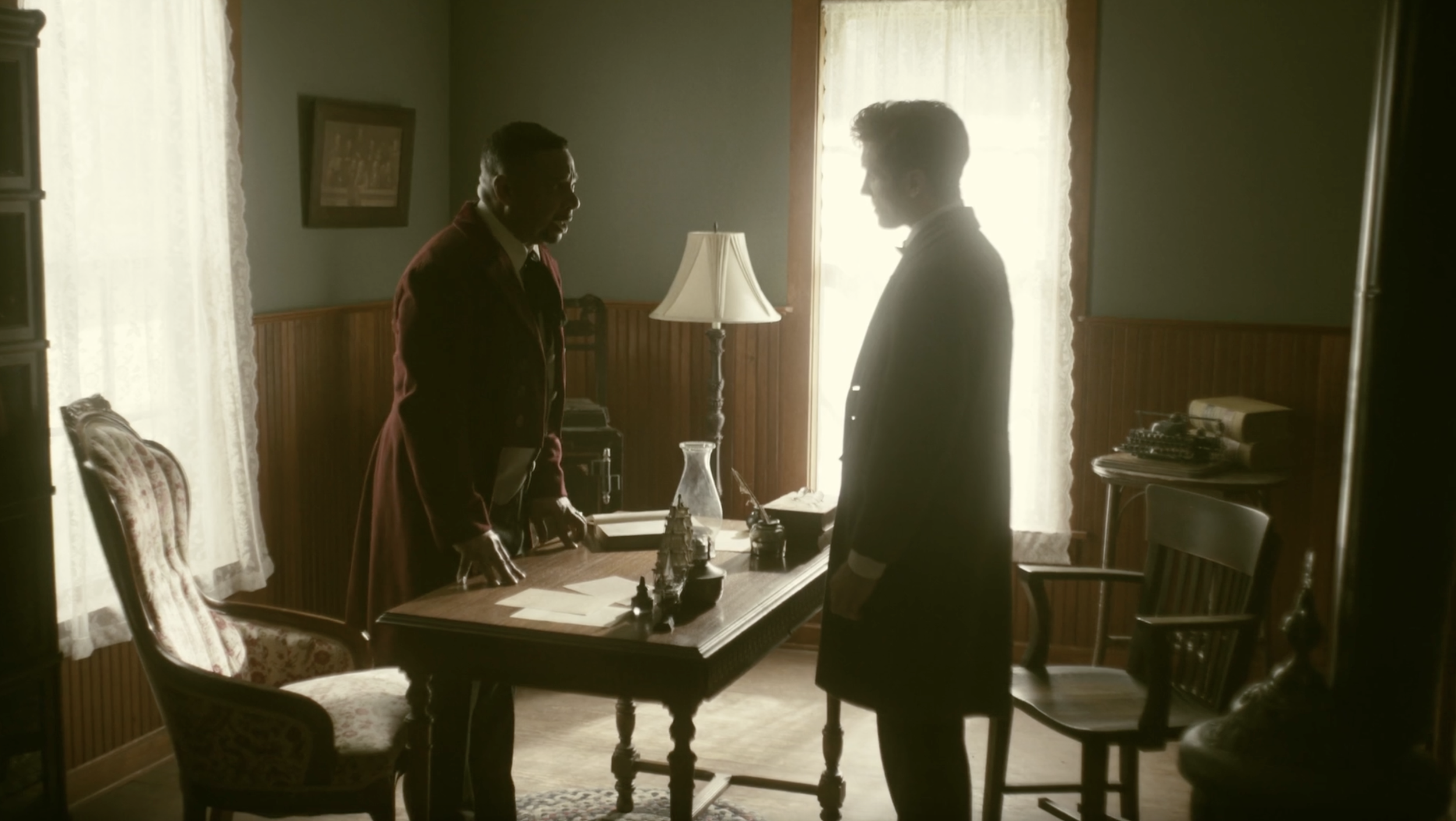 A Black man in a long red vintage coat stands behind a desk in front of a white man in a dark coat.