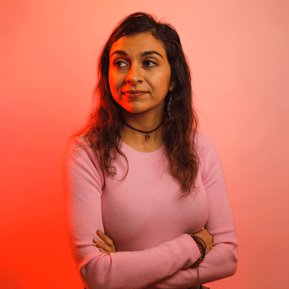 Colette Ghunim a Mexican and Palestinian American woman looks off into the distance. She is wearing a long sleeved pink shirt and is standing in front of a slightly darker pink background.