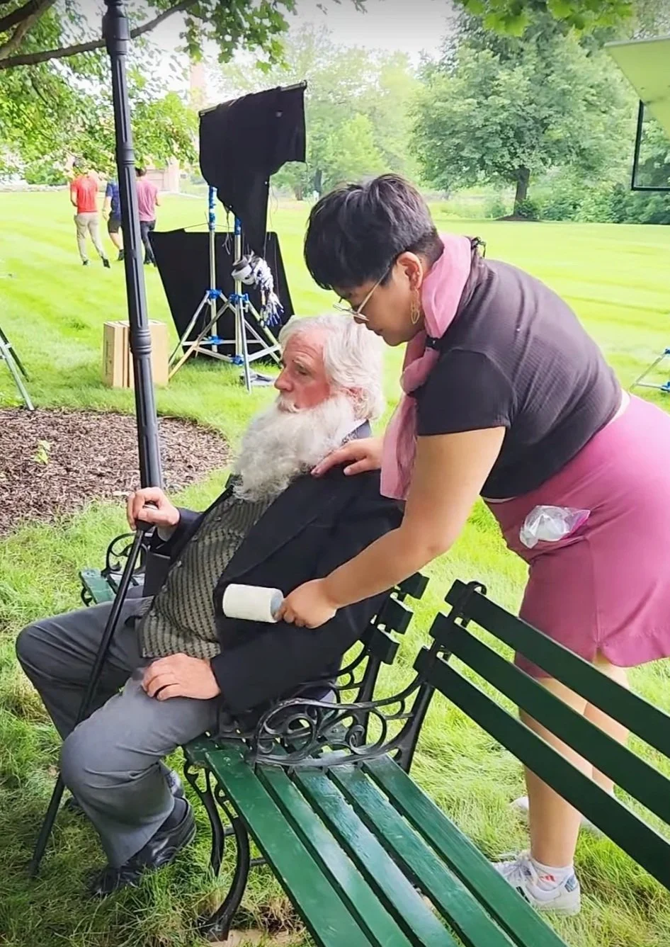 Regina, an Asian American woman uses a lent roller on a older actor sitting down on a park bench.