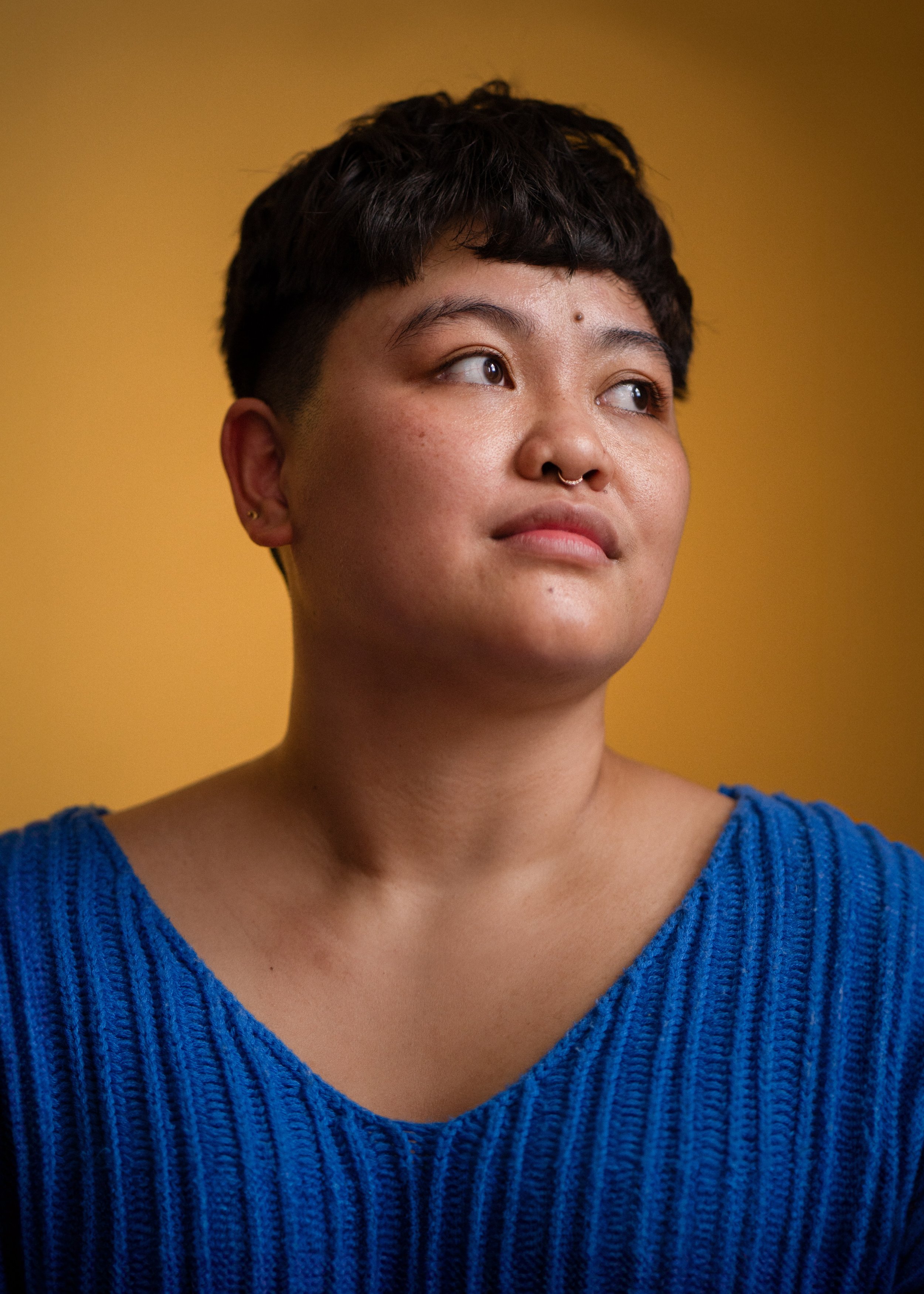Regina, a young Asian American woman with short black hair, wears a bright blue shirt and gazes thoughtfully into the distance. The background is a soft, vignetted mustard yellow.