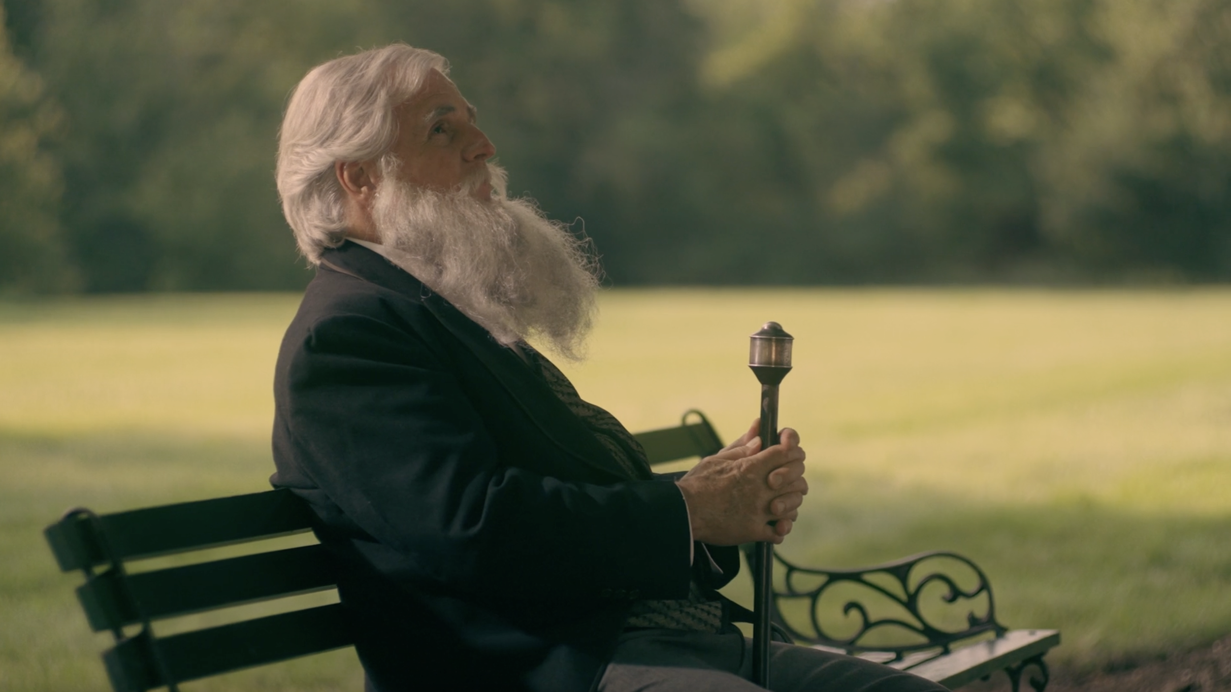An older man with a white hair and long beard sits on a park bench holding a cane.
