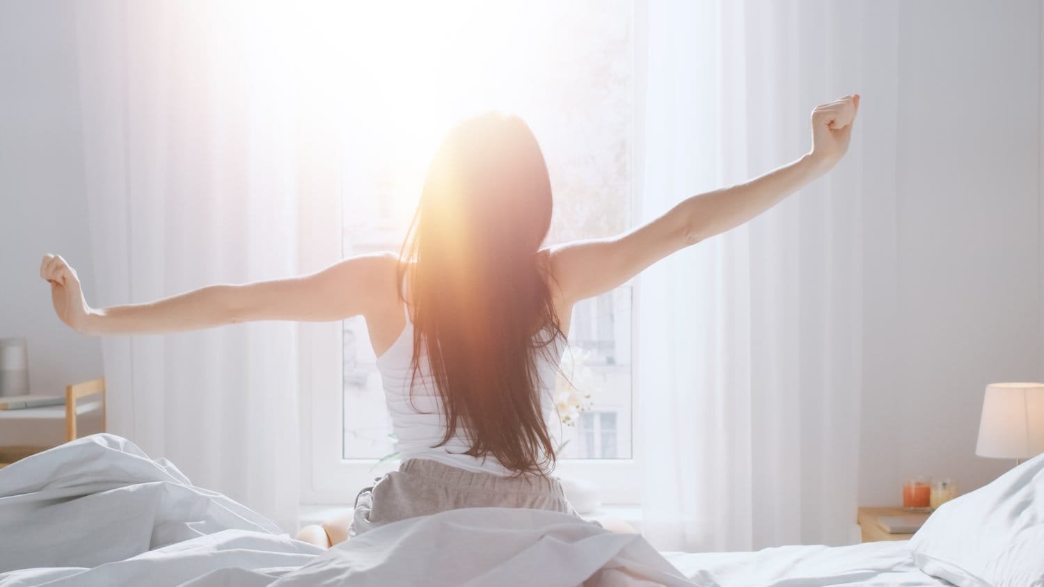 Frau streckt sich morgens im Bett vor hellem Fenster – Symbolbild für erholsamen Schlaf durch bessere Atmung.